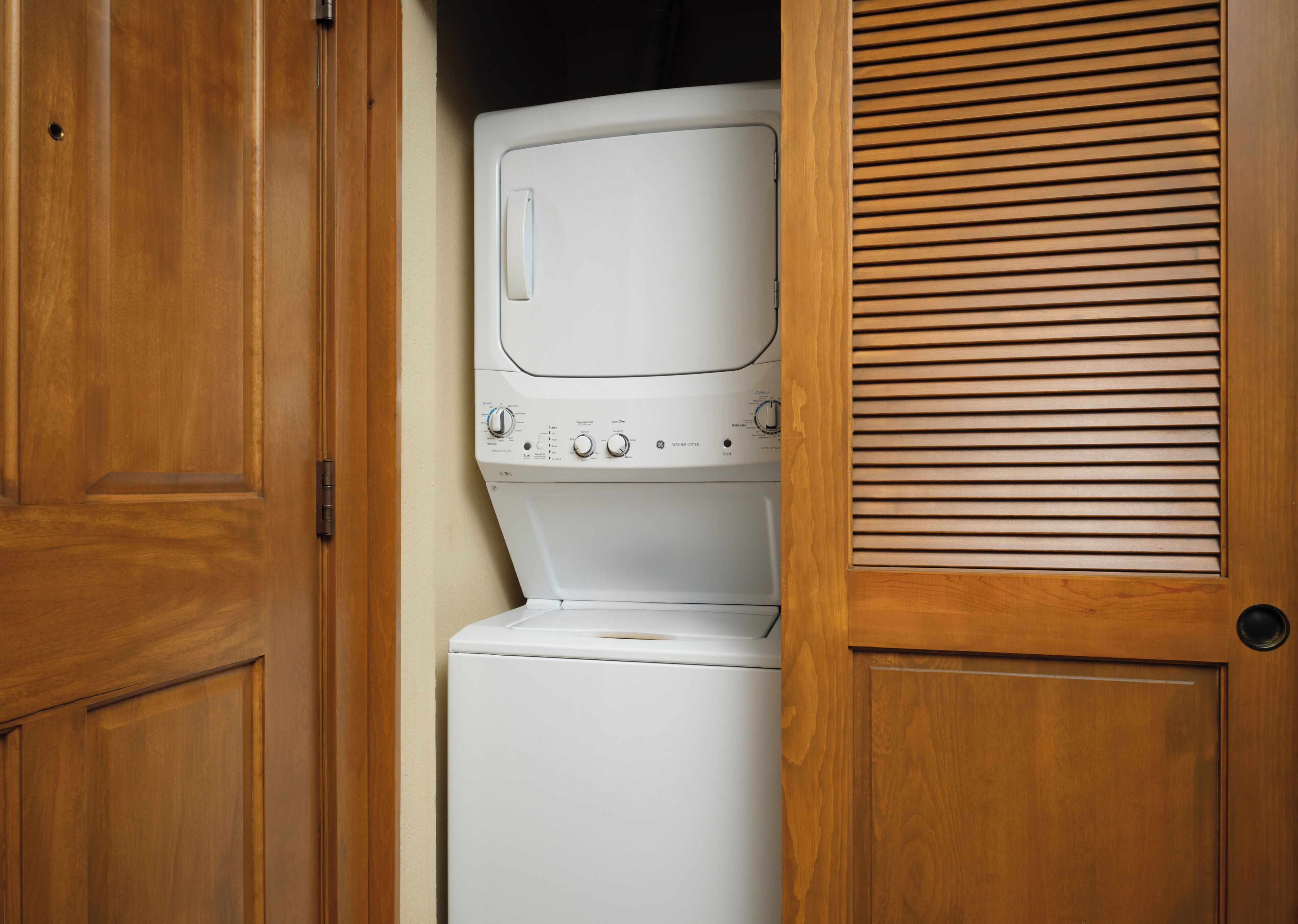 A laundry room with a stacked washer and dryer, wooden cabinets, and a wooden door with a louvered panel.