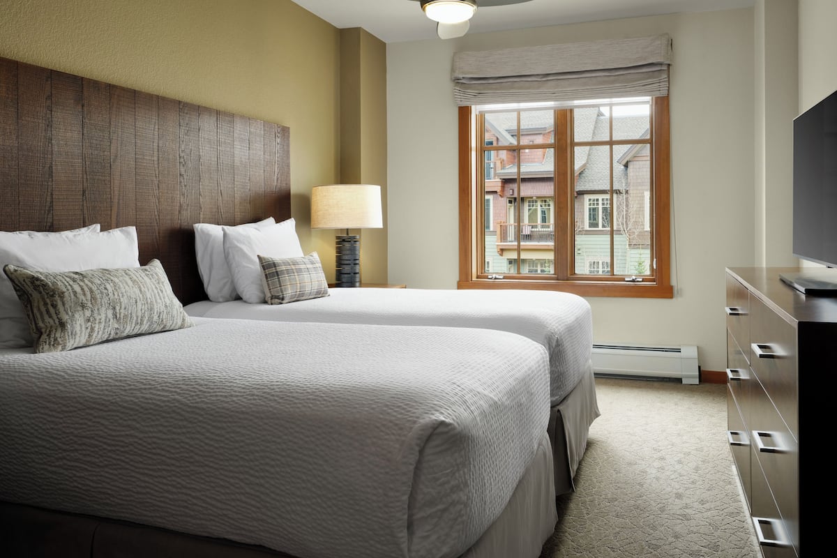 A hotel room with two beds, a ceiling fan, a window with a view of buildings, a lamp, and a TV.
