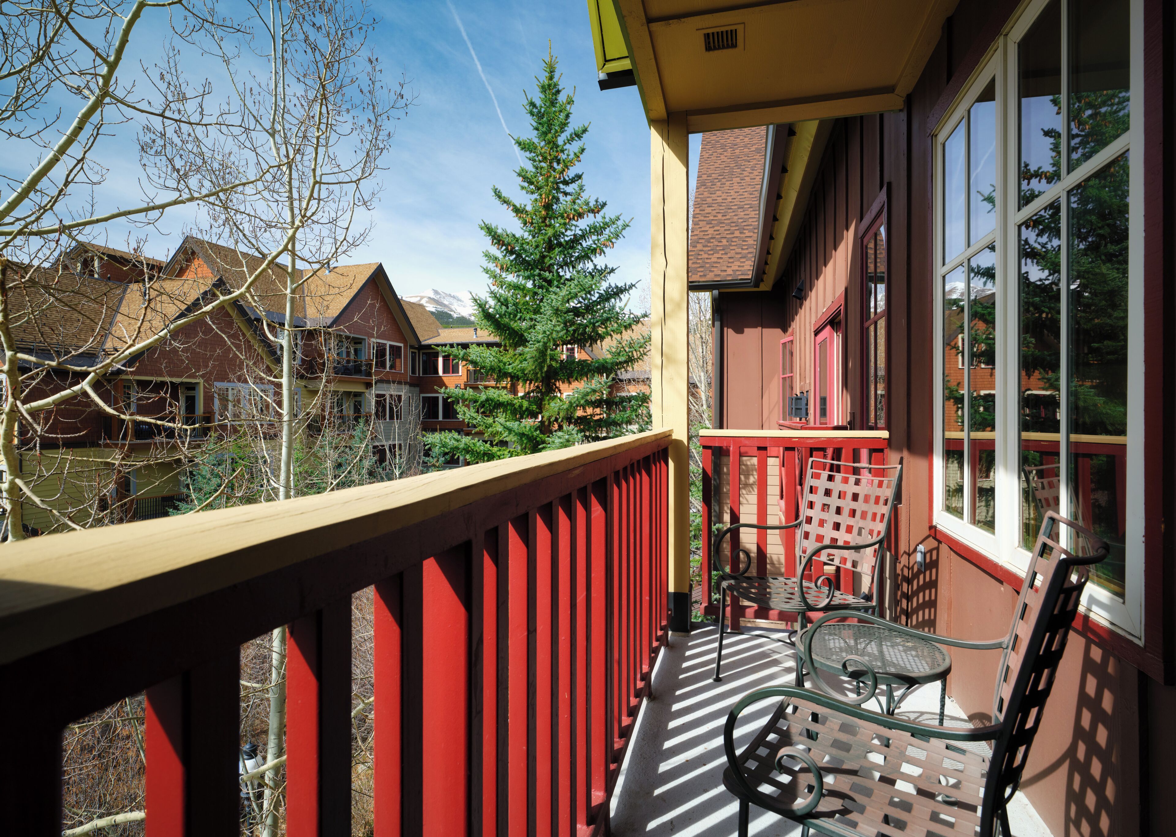 A balcony with a red railing, a glass table, and a chair, overlooking a residential area with houses and a tree.