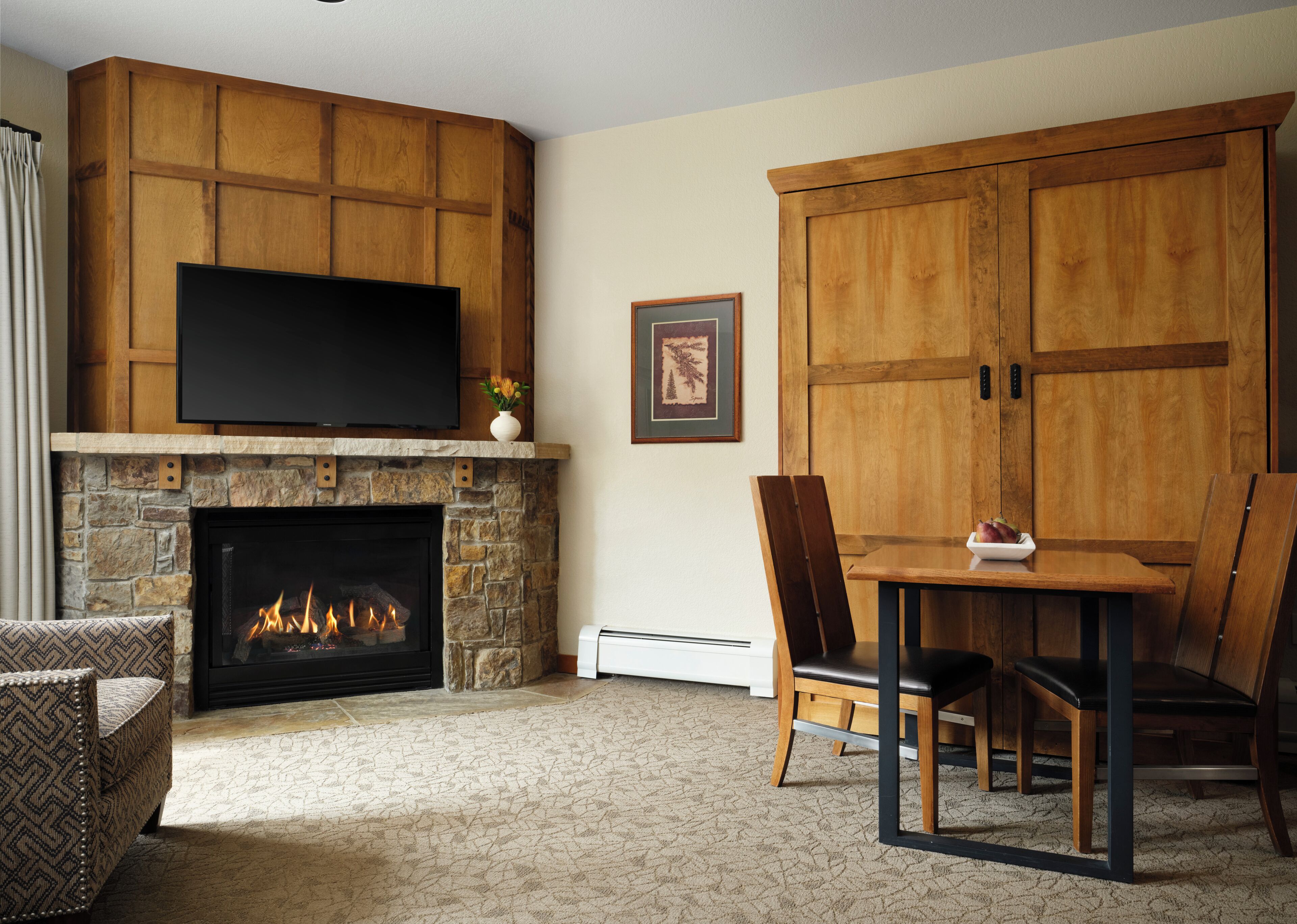 A living room with a stone fireplace, a flat-screen TV mounted above it, a dining table with chairs, and a large wooden cabinet.