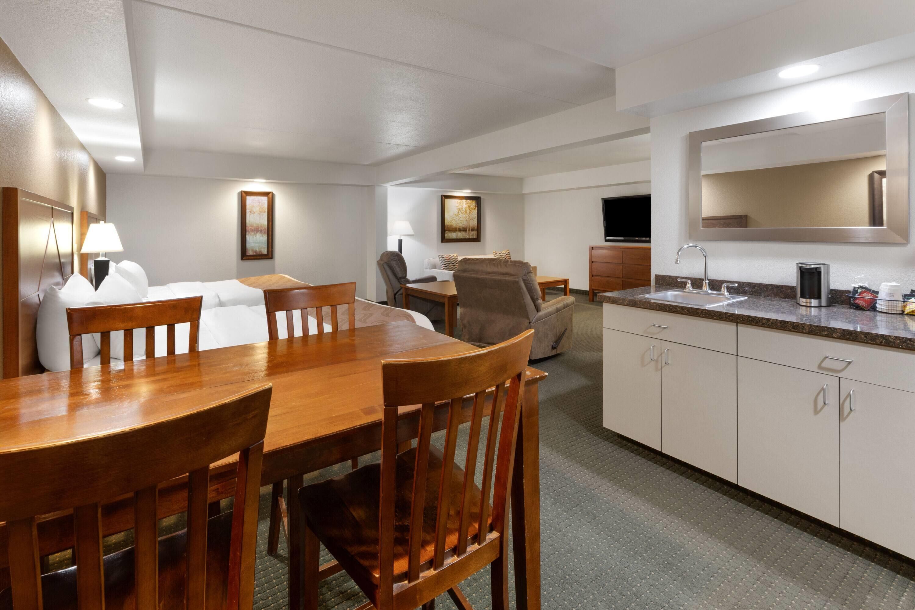 A hotel room with a wooden dining table, chairs, a sofa, a TV, a kitchen area with a sink and cabinets, and a large mirror.