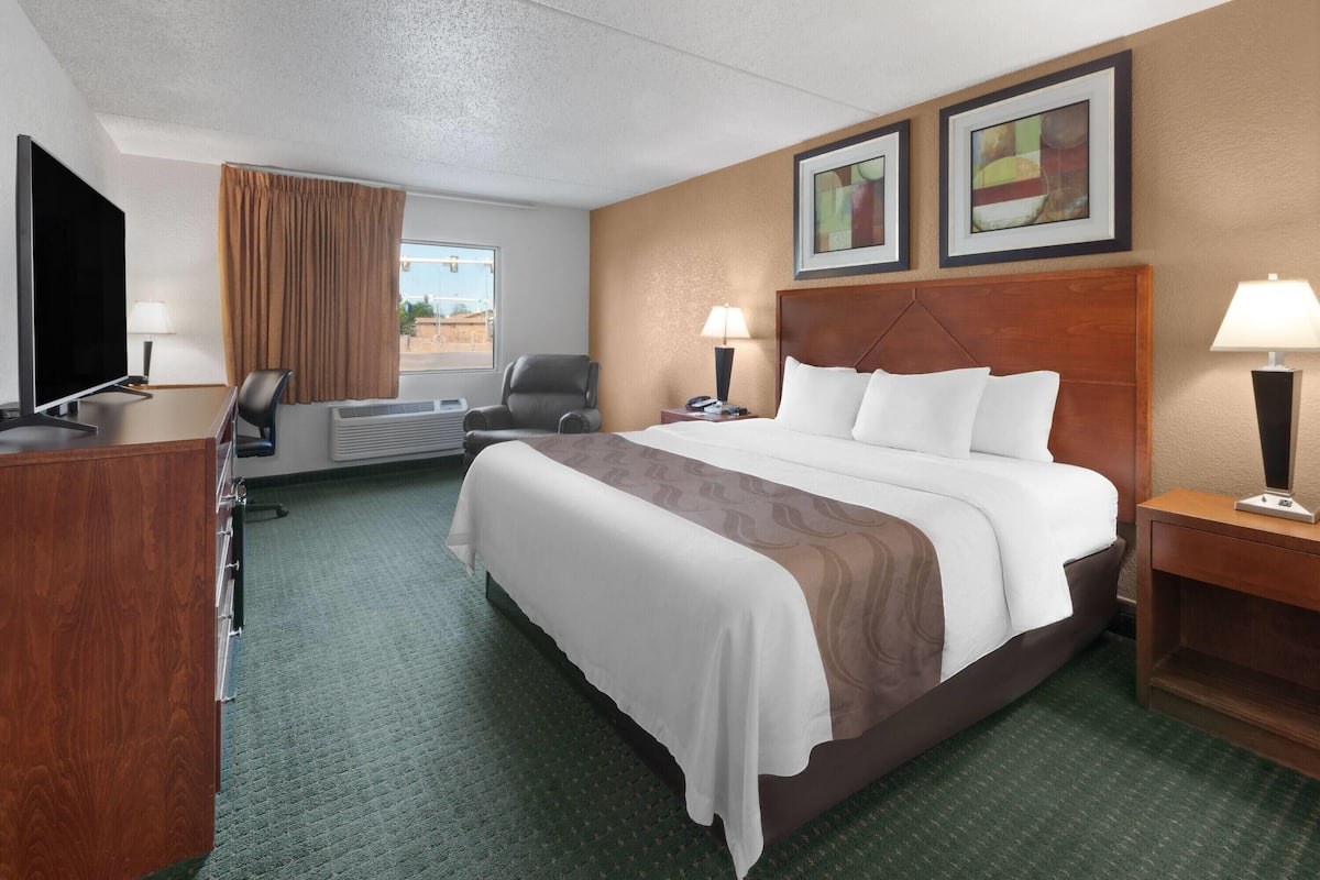 A hotel room with a large bed, a desk with a flat-screen TV, a chair, a desk lamp, and two framed pictures on the wall.