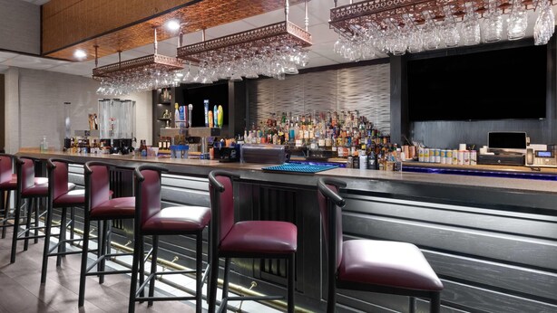 Zona de bar con varios taburetes rojos, una barra con varias botellas y una caja registradora, y una pantalla grande sobre la barra.