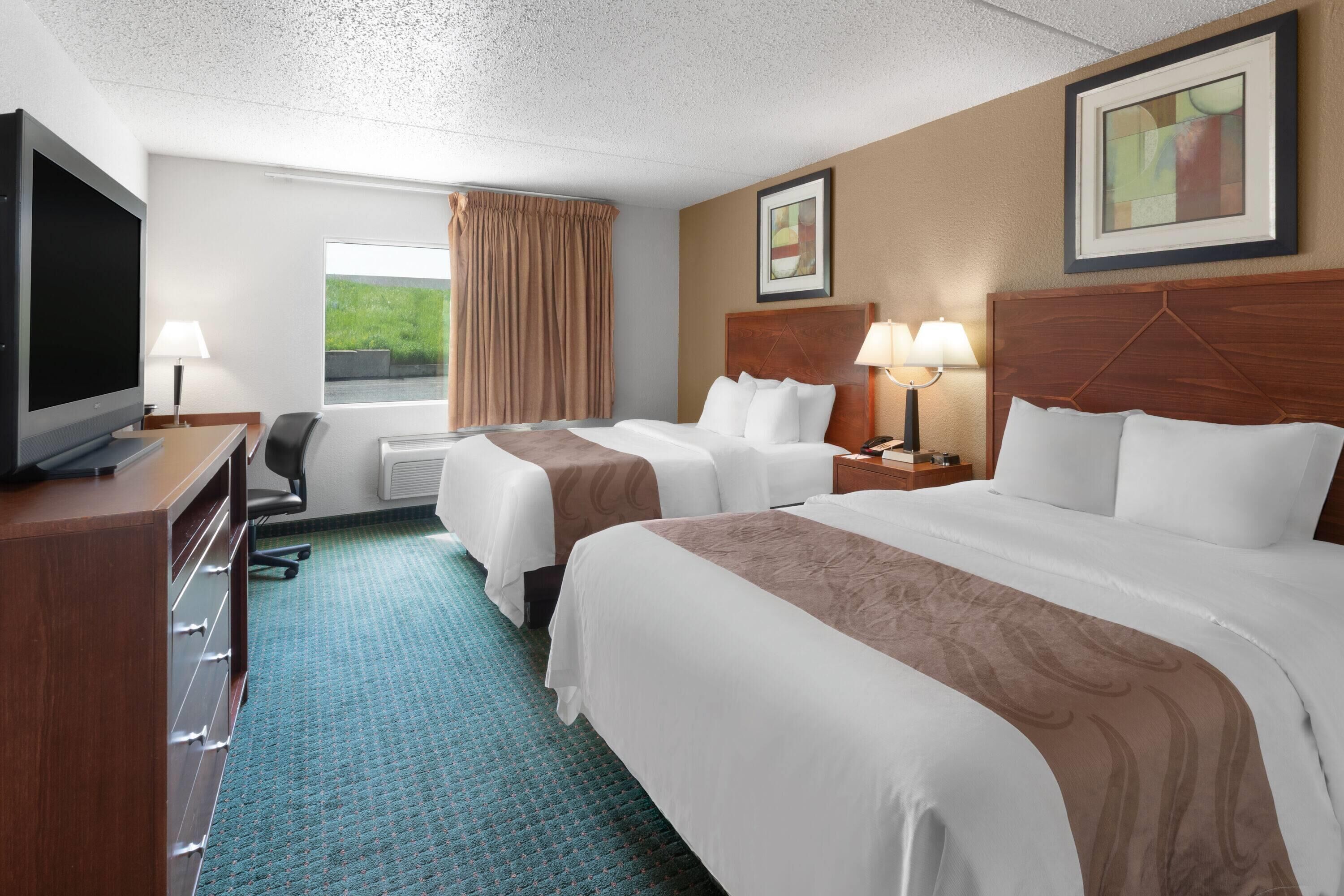 A hotel room with two beds, a TV, a desk, and a window with curtains.