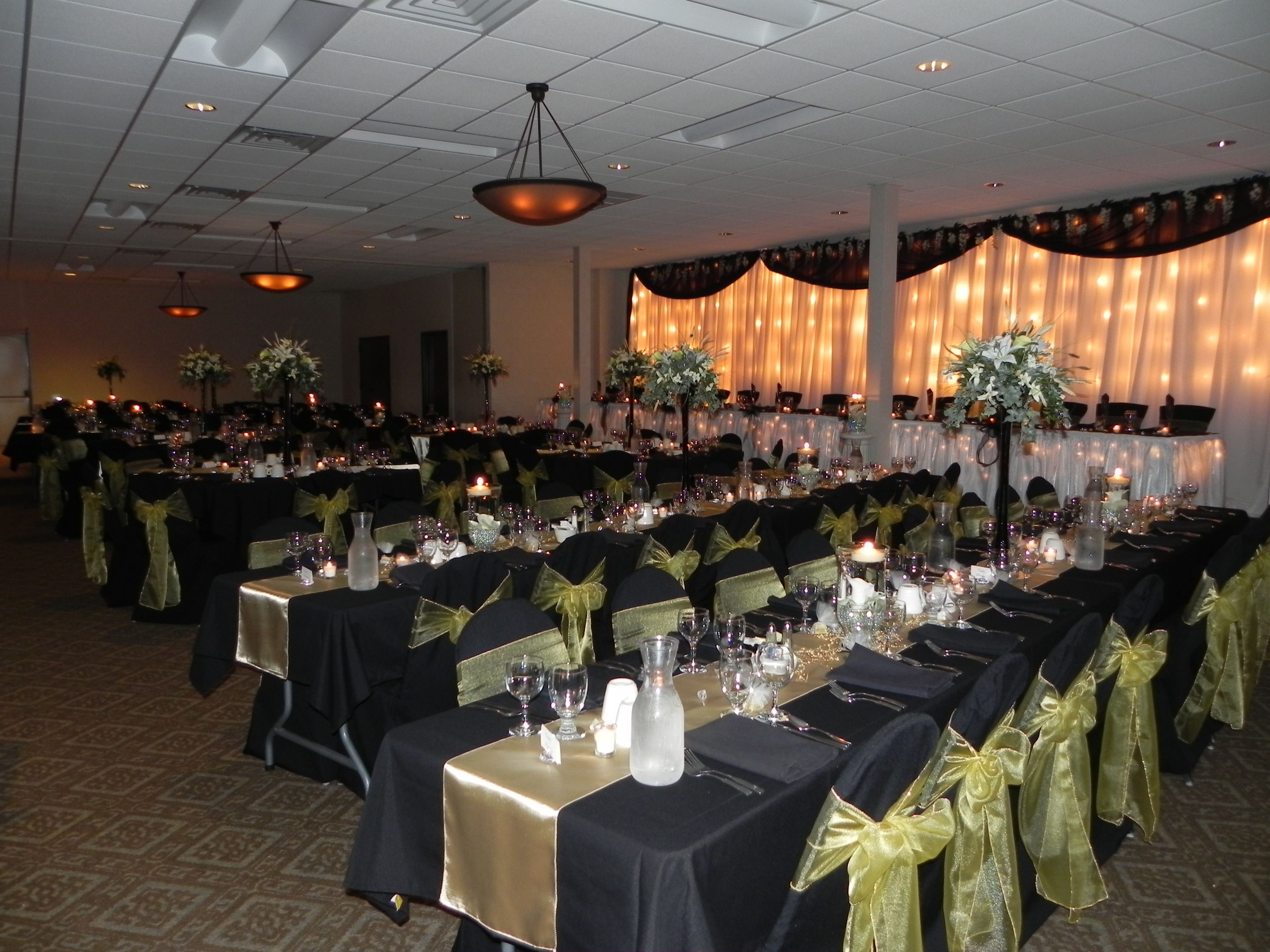 Une salle de banquet avec de longues tables dressées pour un événement formel, agrémentée de nappes noires, de rubans dorés et de compositions florales en guise de centre de table.