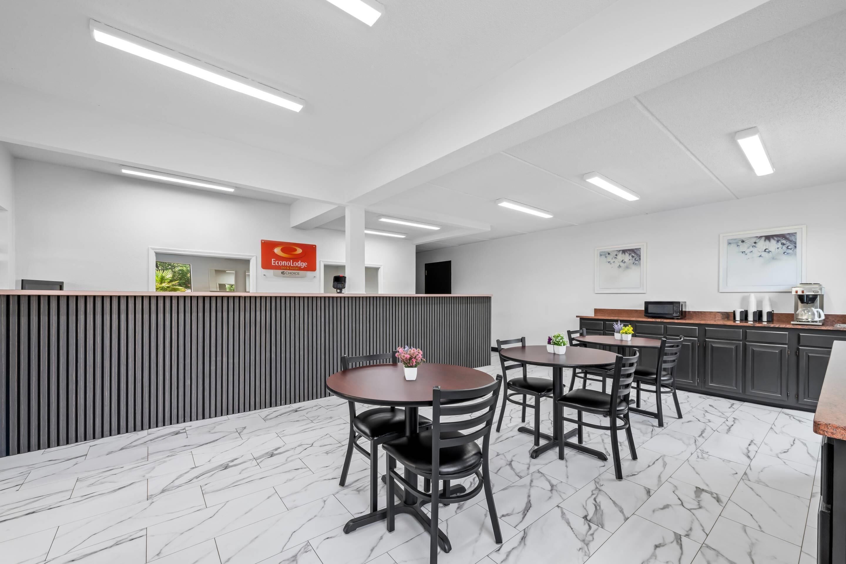 A modern hotel lobby with a reception desk, a dining area with marble flooring, and a kitchen area with dark cabinetry.