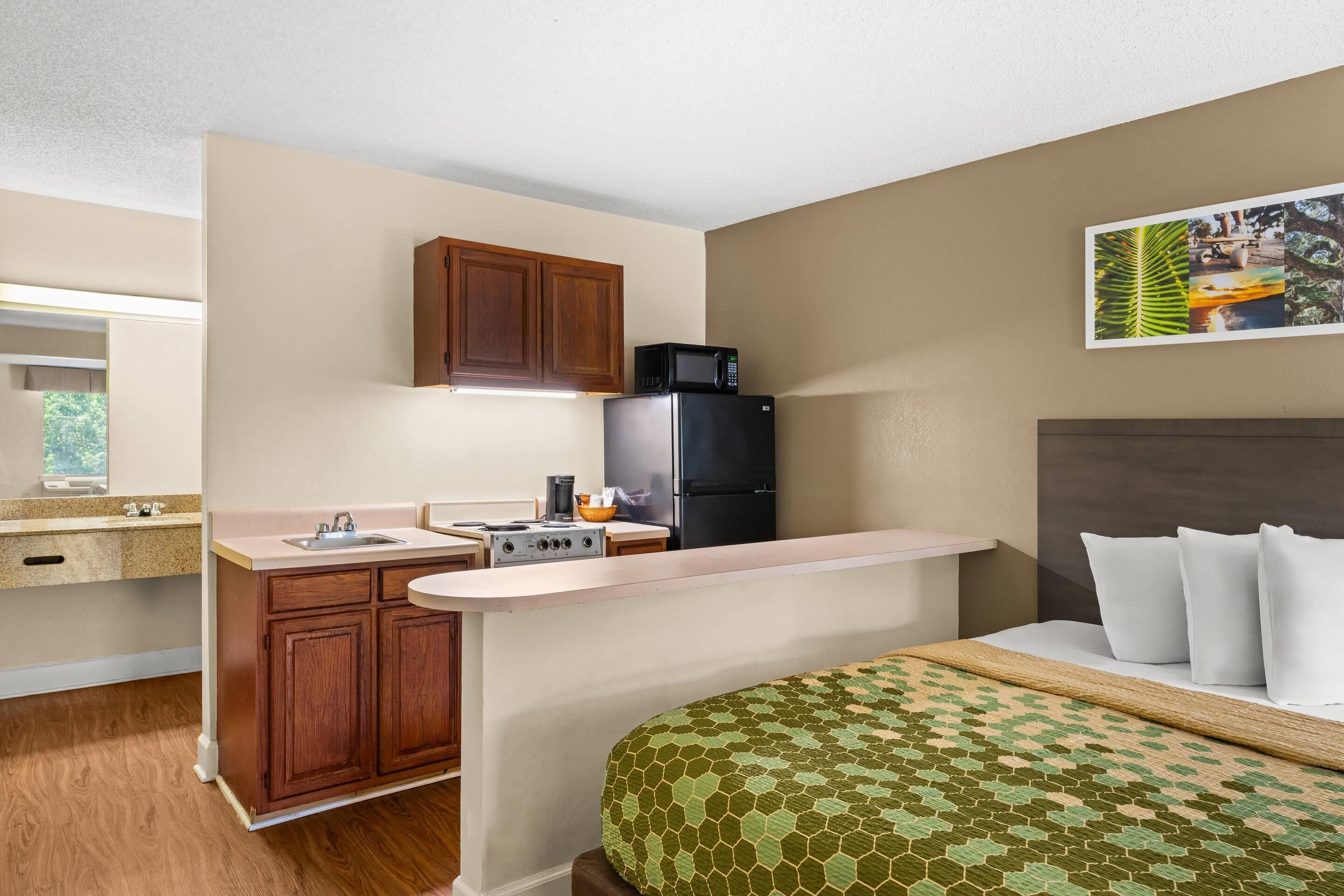 A compact hotel room with a kitchenette, a bed with white pillows, and a wall-mounted artwork.