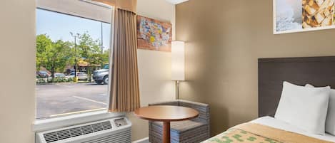 A hotel room with a bed, a round table, a chair, a window with curtains, and a wall-mounted lamp.