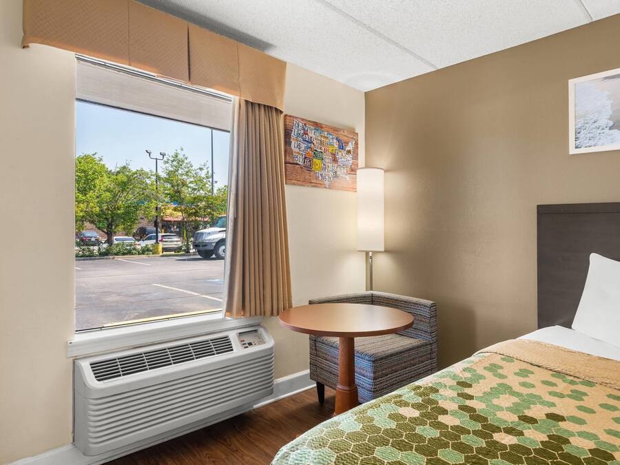 A hotel room with a bed, a round table, a chair, a window with curtains, and a wall-mounted lamp.