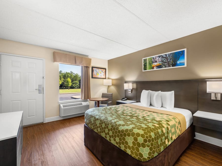 Habitación de hotel con cama, escritorio, silla, ventana con vistas a zonas verdes y un cuadro colgado en la pared.