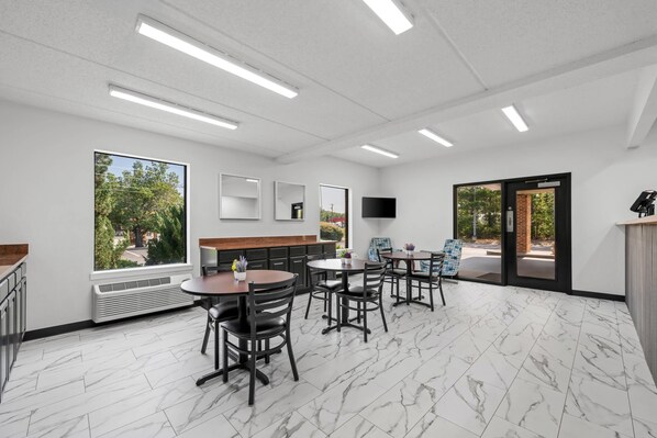 A modern interior with a marble floor, a dining area with tables and chairs, a large window with a view of trees, and a flat-screen TV mounted on the wall.