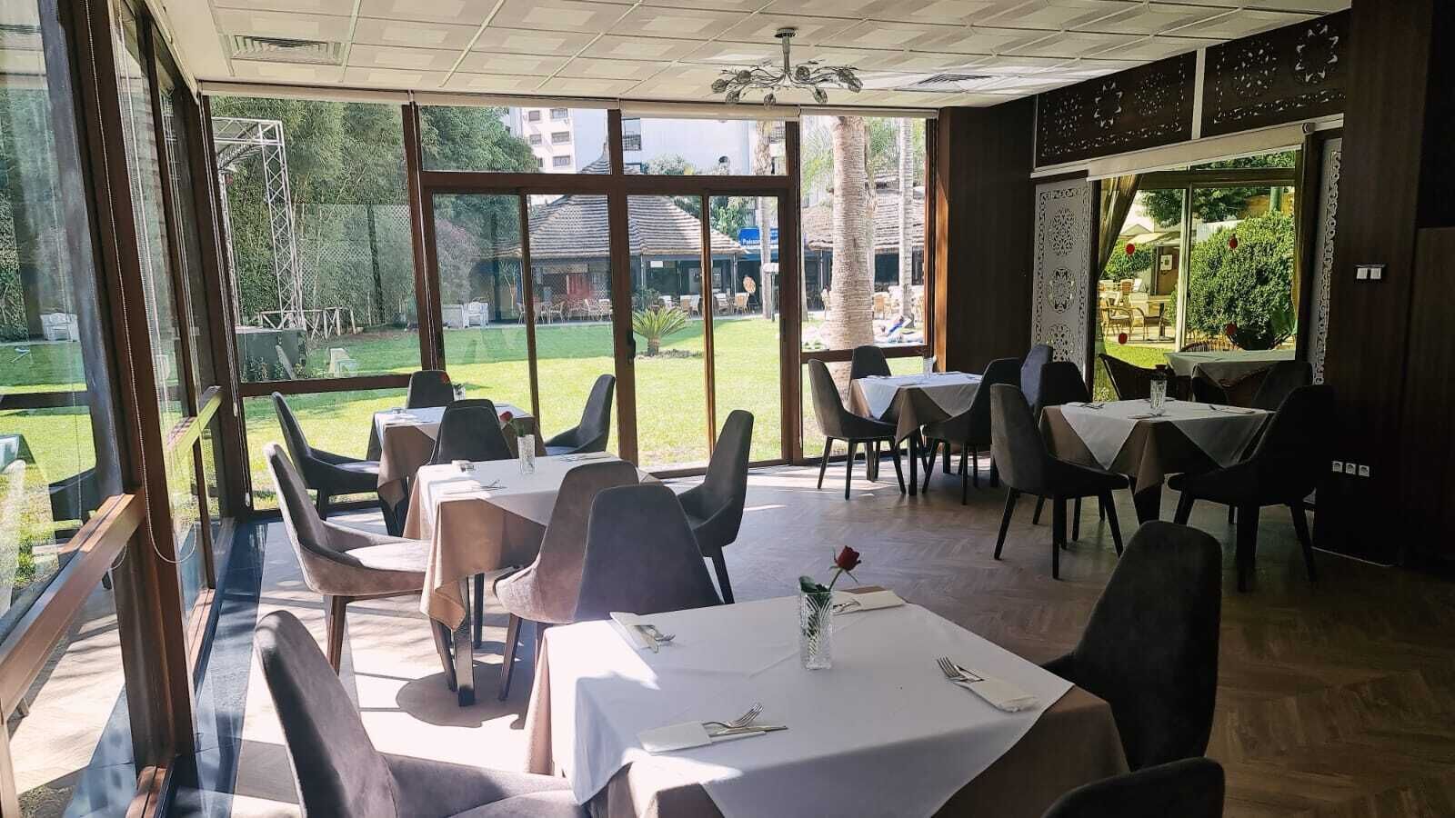 Een restaurant met tafels en stoelen voor diners, grote ramen met uitzicht op de omgeving, en een plafond met een decoratieve verlichtingsarmatuur.
