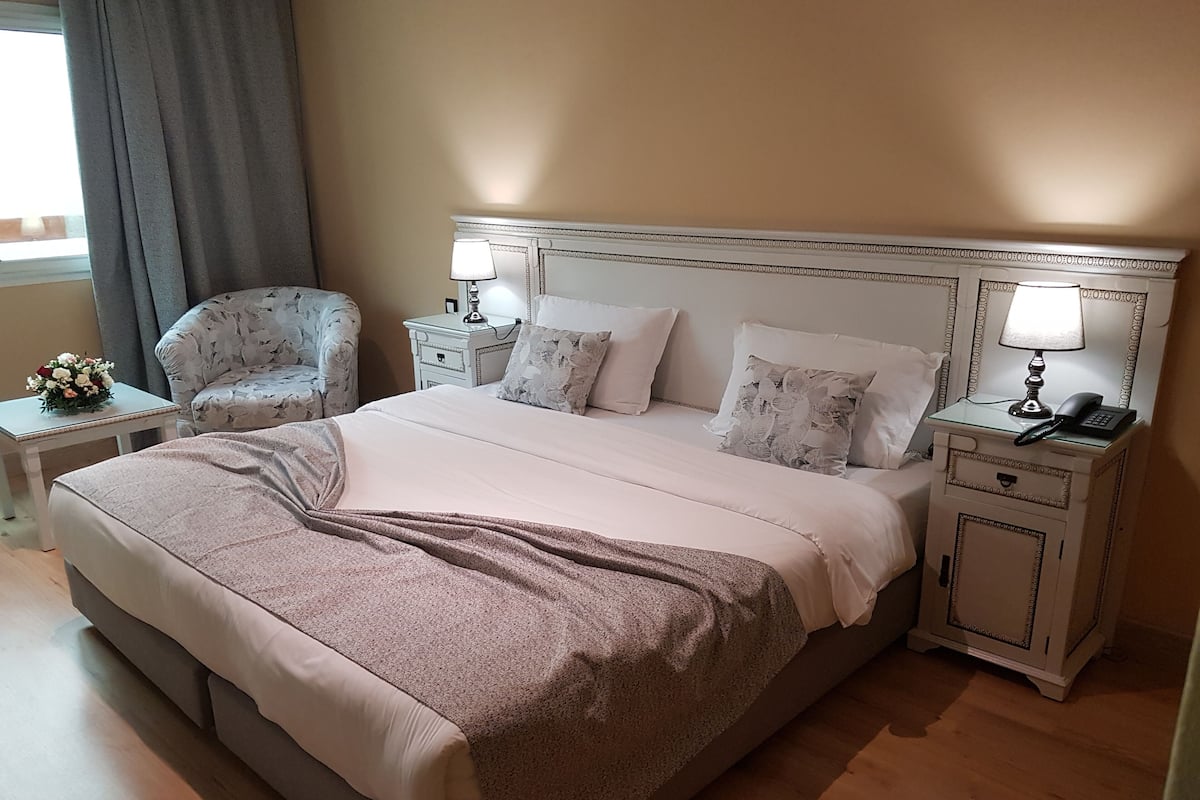 A neatly made bed with a patterned headboard, two bedside tables with lamps, a floral arrangement, and a chair.