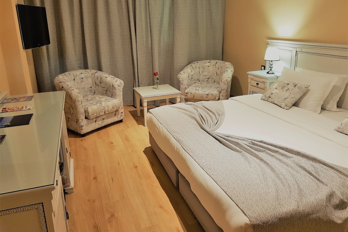 A hotel room with a bed, two armchairs, a small table with a vase, and a nightstand with a lamp.
