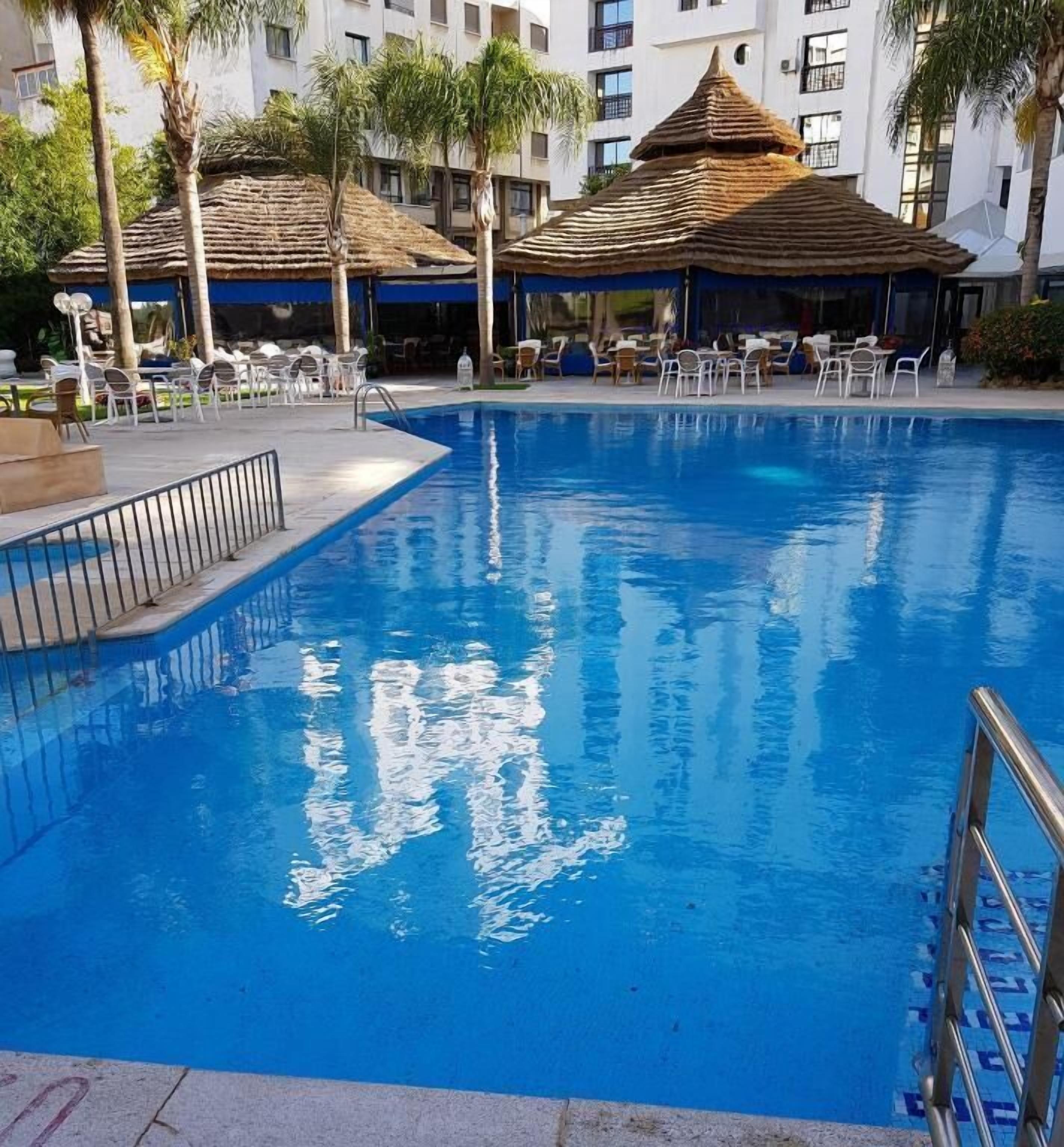 A swimming pool with lounge chairs and umbrellas, surrounded by palm trees and a building with multiple windows.