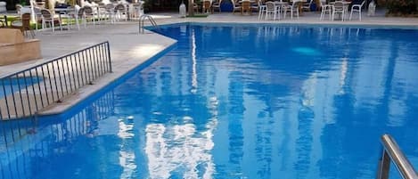 Una piscina con sillas de descanso y sombrillas, rodeada de palmeras y un edificio con numerosas ventanas.