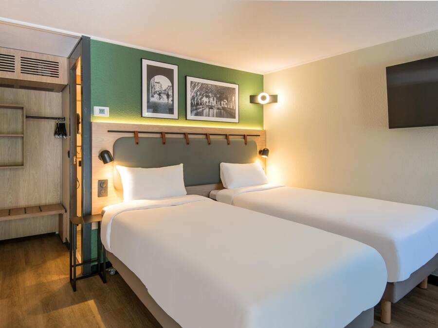 Ein Hotelzimmer mit zwei Einzelbetten, einem Holzschrank, einer kleinen Sitzecke und einem an der Wand befestigten Fernseher.