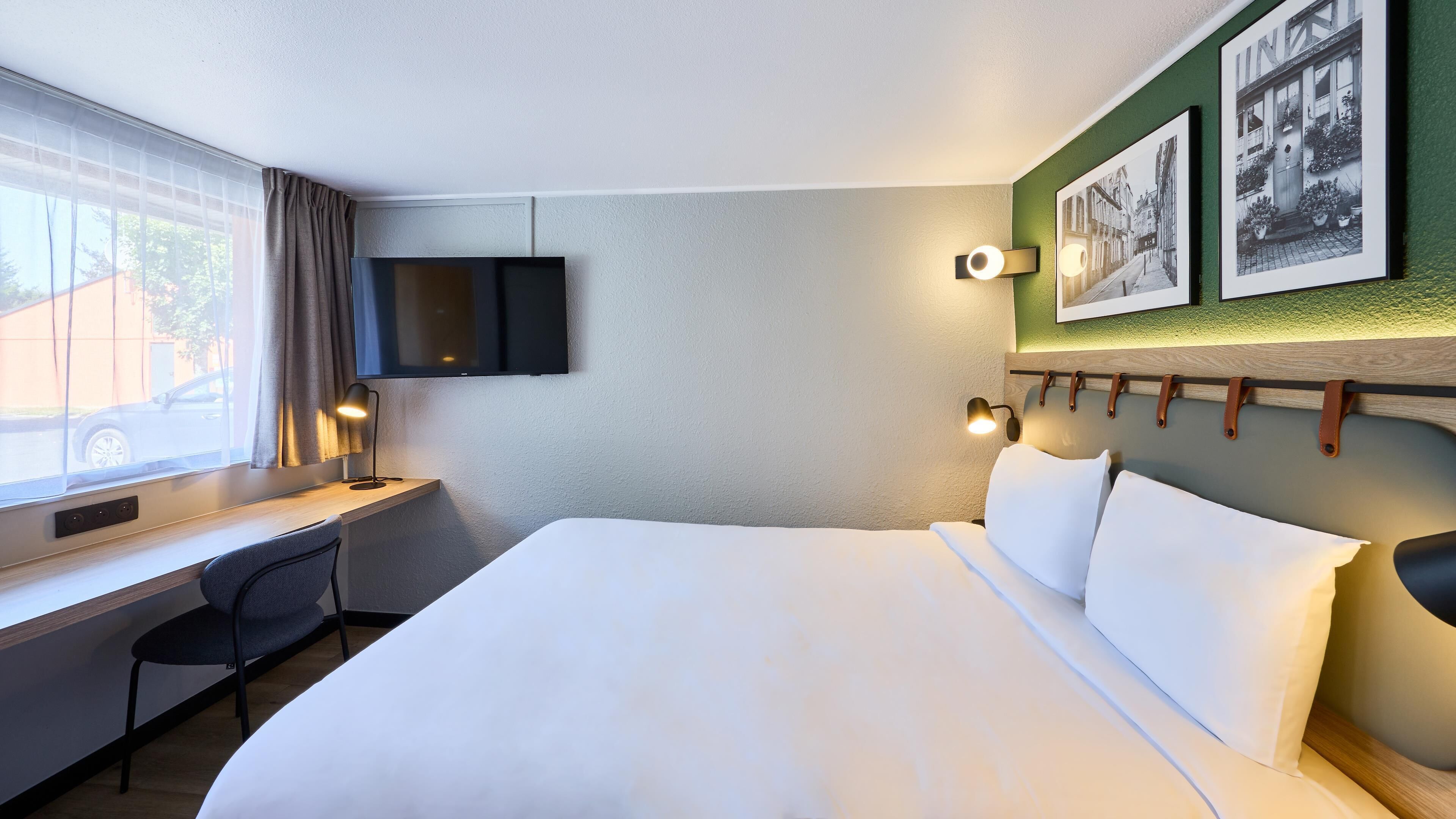 A hotel room with a bed, desk, chair, TV, and wall-mounted lights.