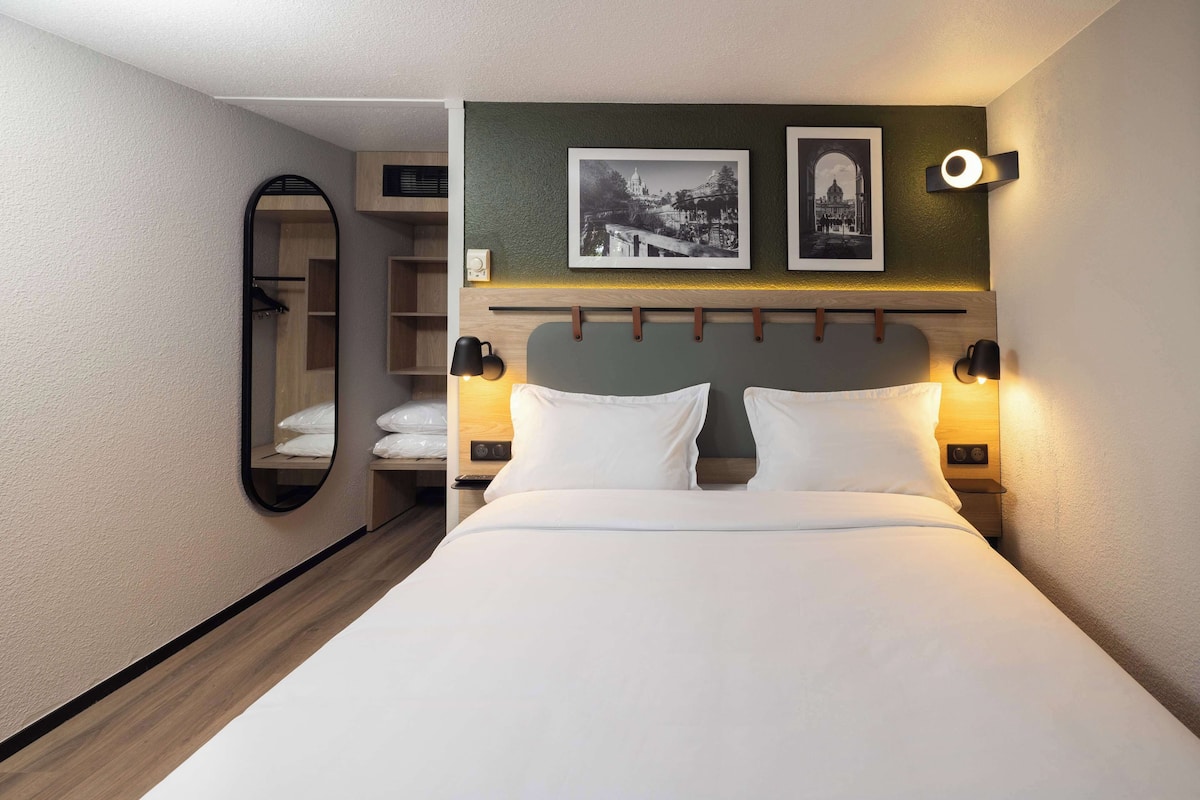 A hotel room with a bed, two bedside lamps, a mirror, and two framed pictures on the wall.
