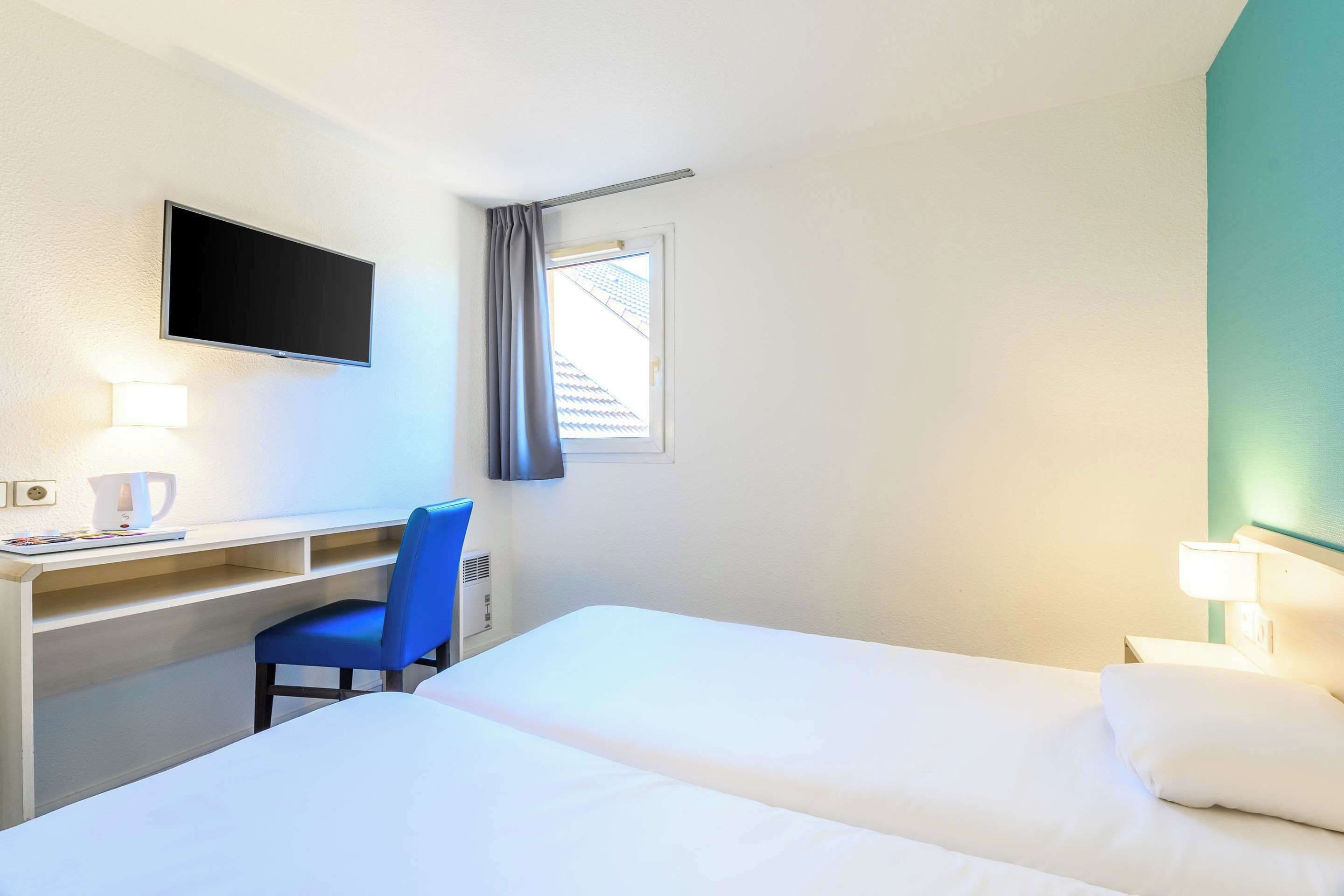 Una habitación de hotel con cama, escritorio, silla, televisor y ventana con cortinas.