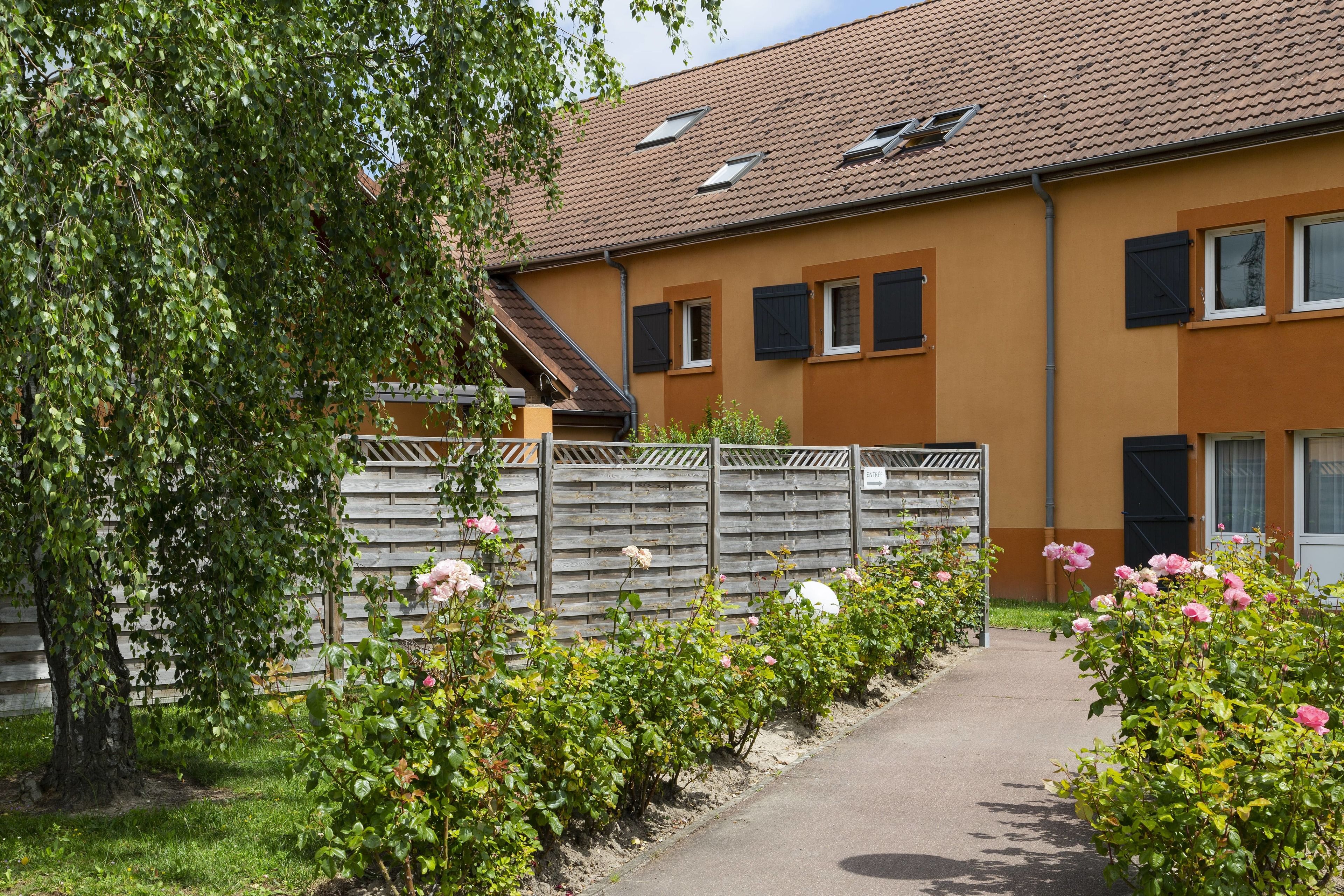 A residential building with a brown facade, multiple windows with shutters, a visible staircase, and a pathway lined with flowering bushes.