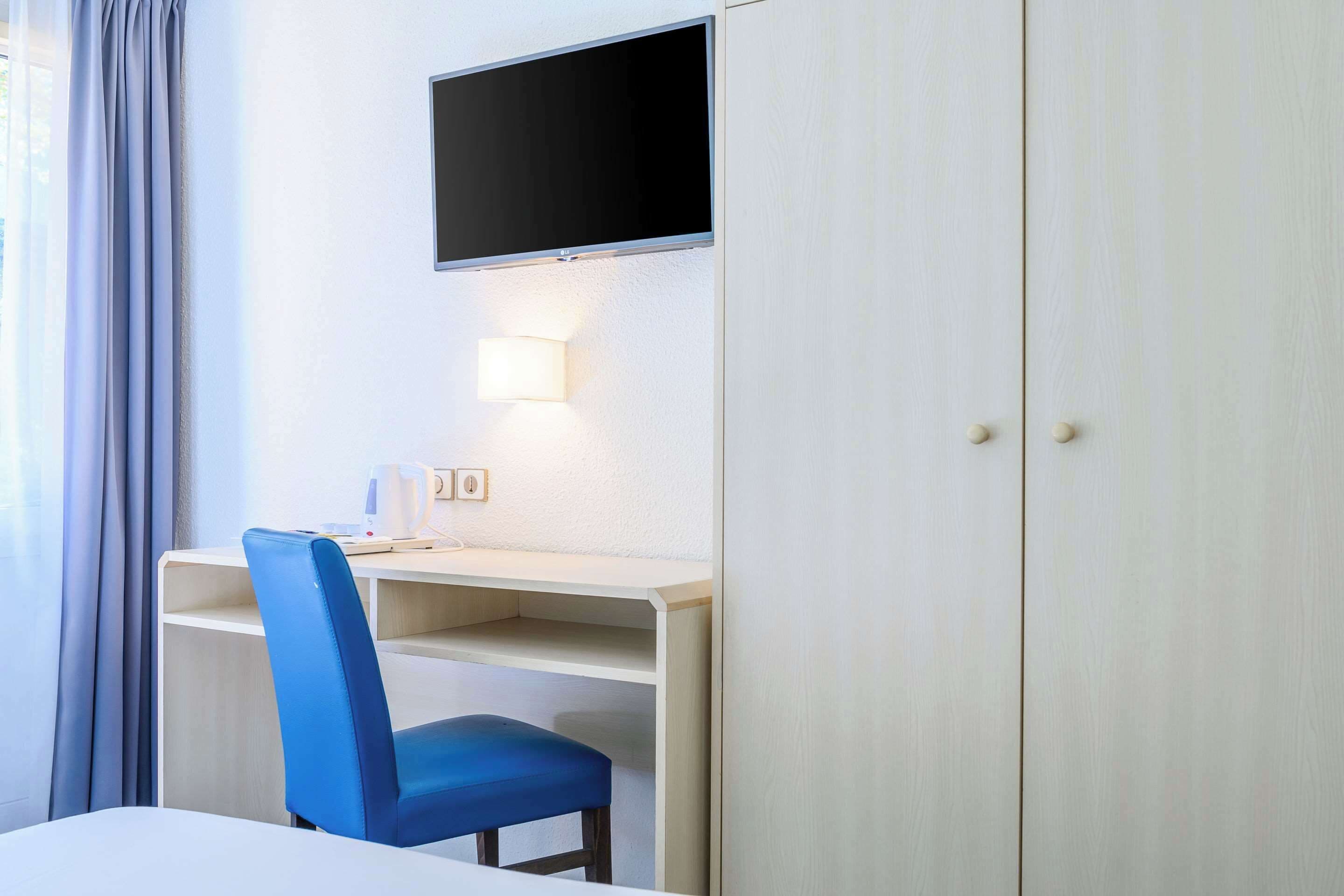 A hotel room with a desk, a chair, a television mounted on the wall, and a wardrobe.