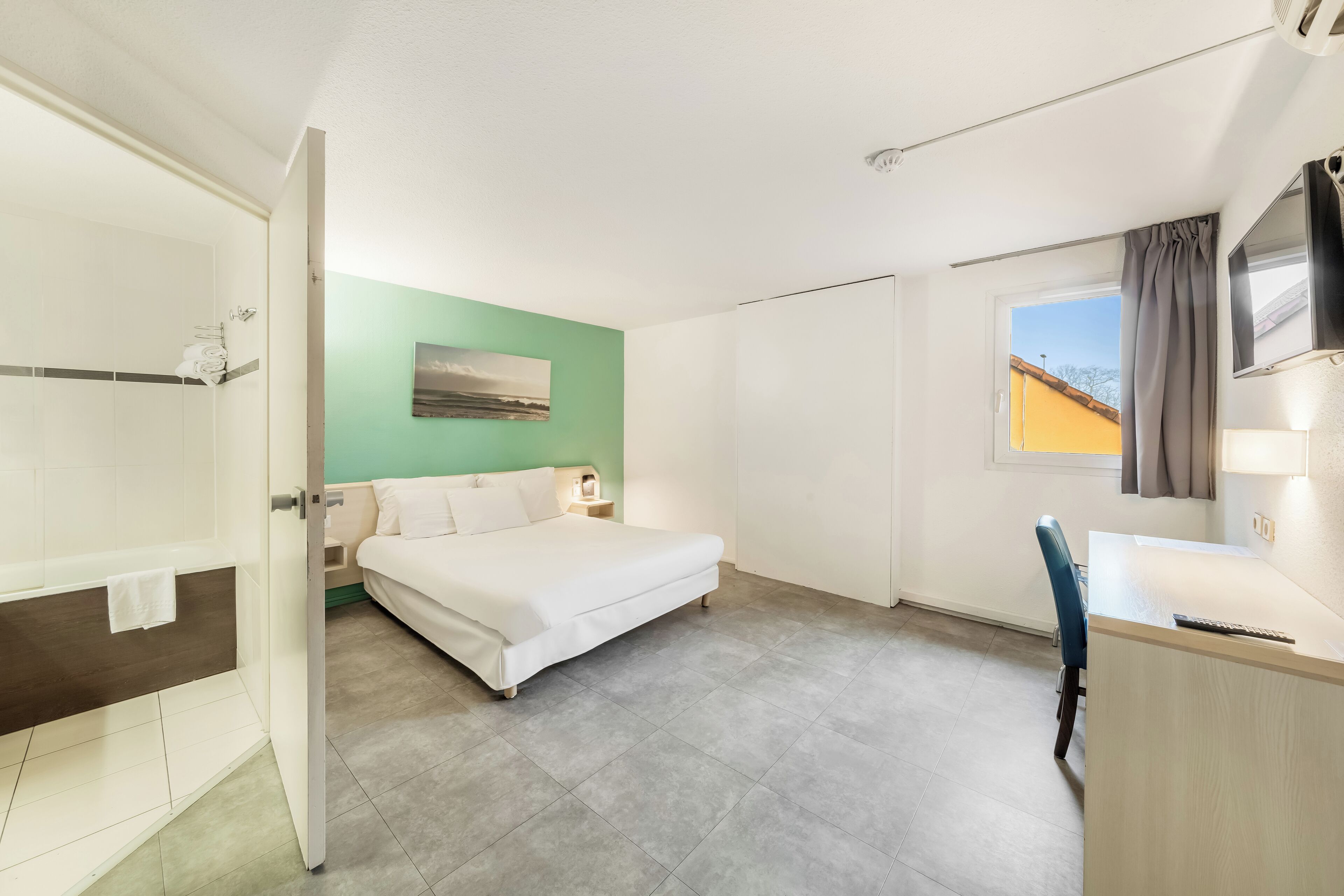 Habitación de hotel con cama, escritorio, silla, zona de ducha y ventana con vistas al exterior.