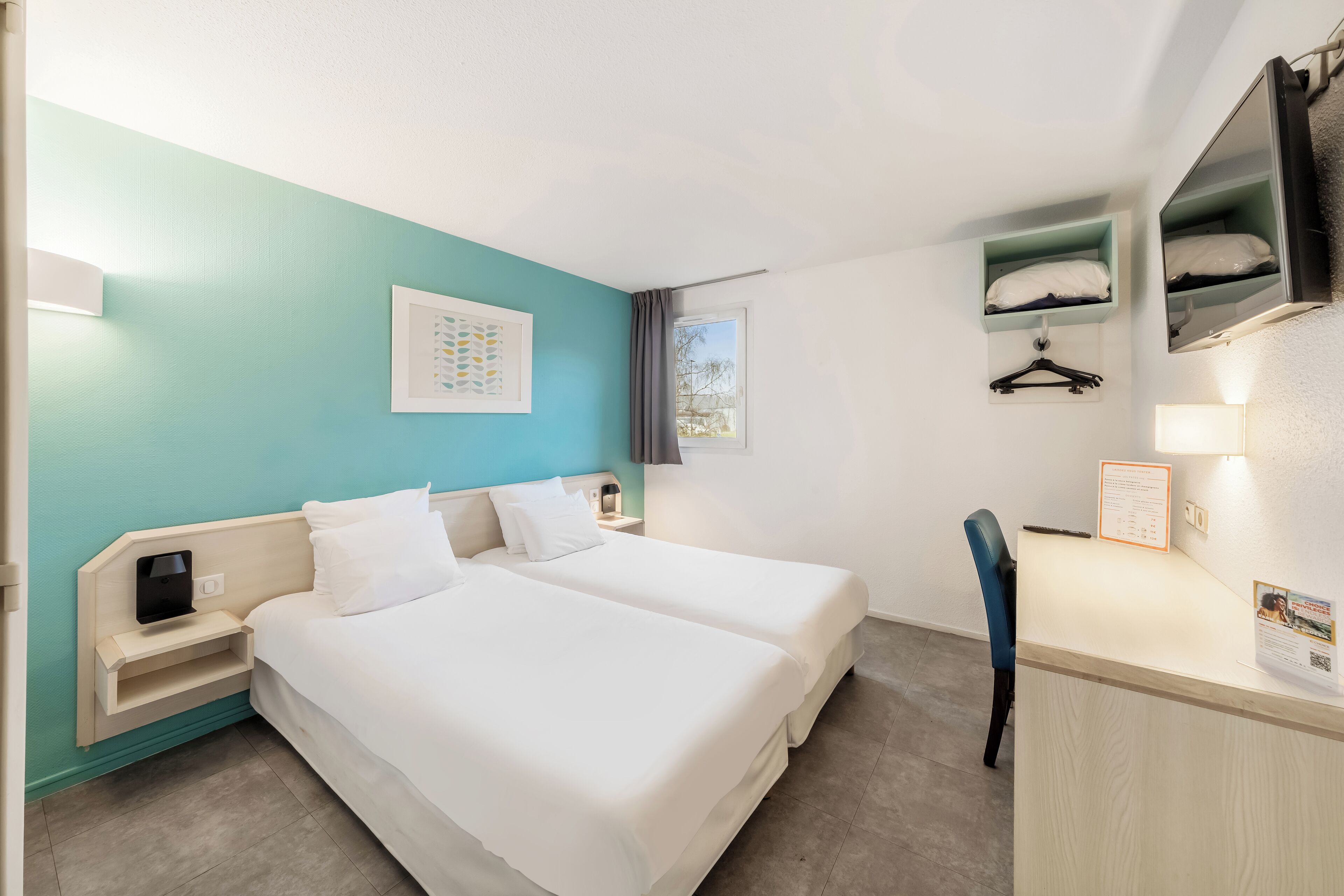 Habitación de hotel con dos camas, un escritorio, una silla, un estante pequeño con perchero y una ventana con cortinas.
