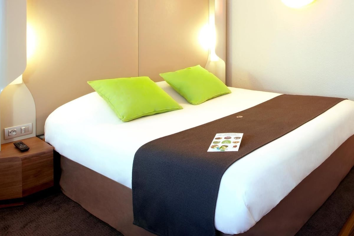 Ein ordentlich bezogenes Bett mit zwei Kissen, ein Nachttisch mit Fernbedienung und eine an der Wand befestigte Leuchte.