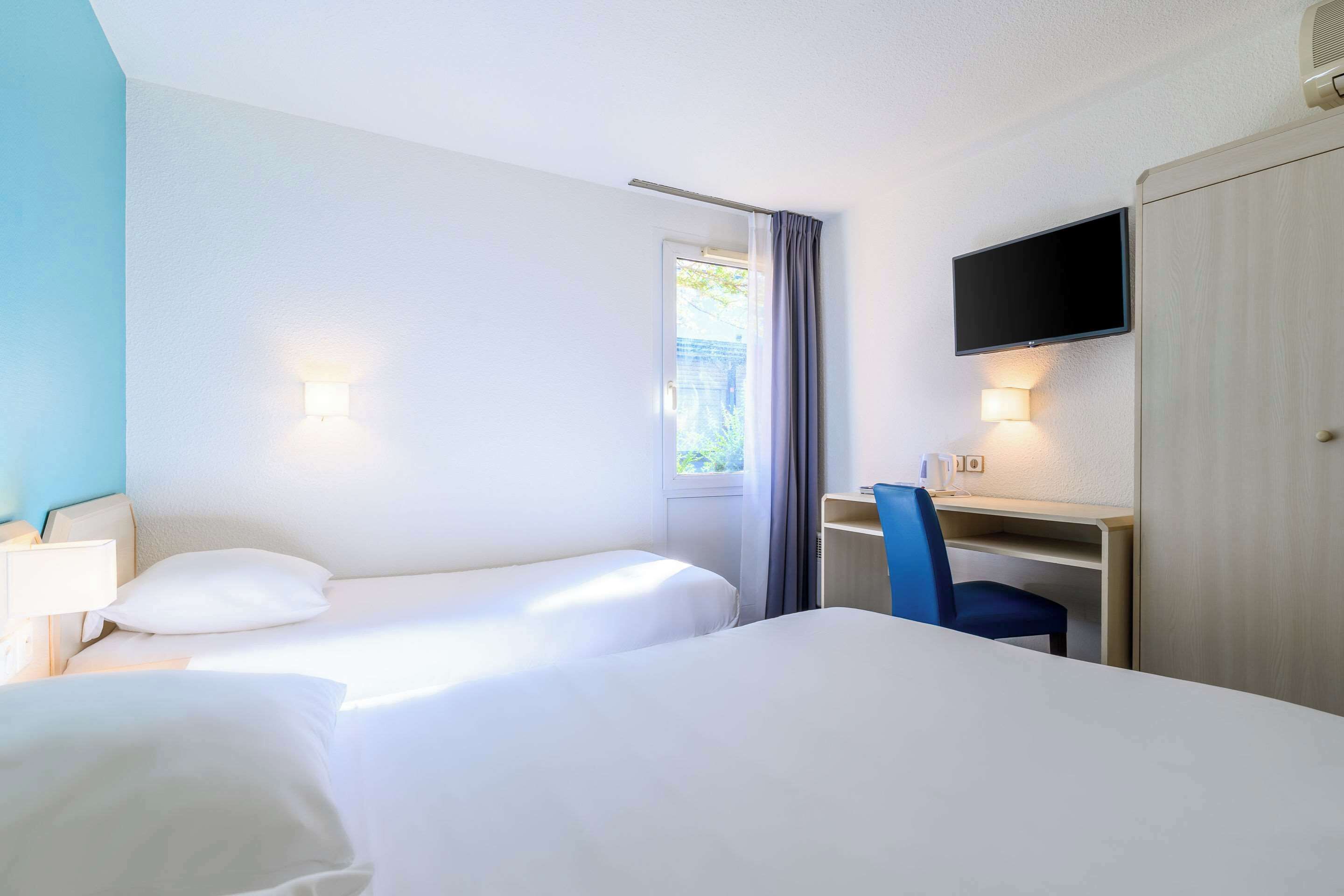 Une chambre d’hôtel avec deux lits, un bureau avec une chaise, une télévision et une fenêtre avec des rideaux.