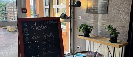 A hotel reception area with a chalkboard displaying room rates and amenities, a small table with magazines, and a view of the outdoor area through the glass doors.