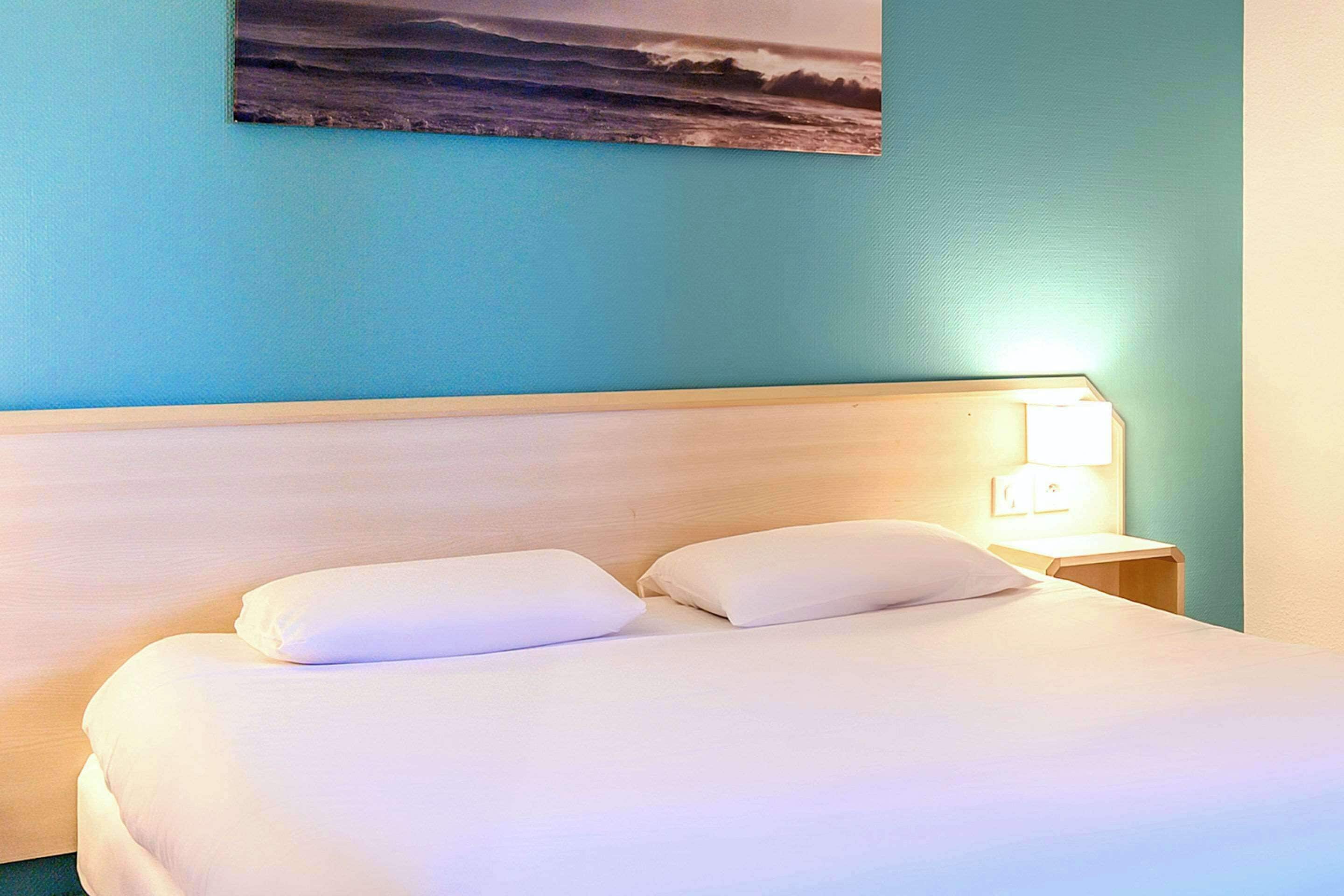 Una cama bien tendida con sábanas y almohadas blancas, cabecera de madera y un cuadro colgado en la pared que representa un paisaje marino.