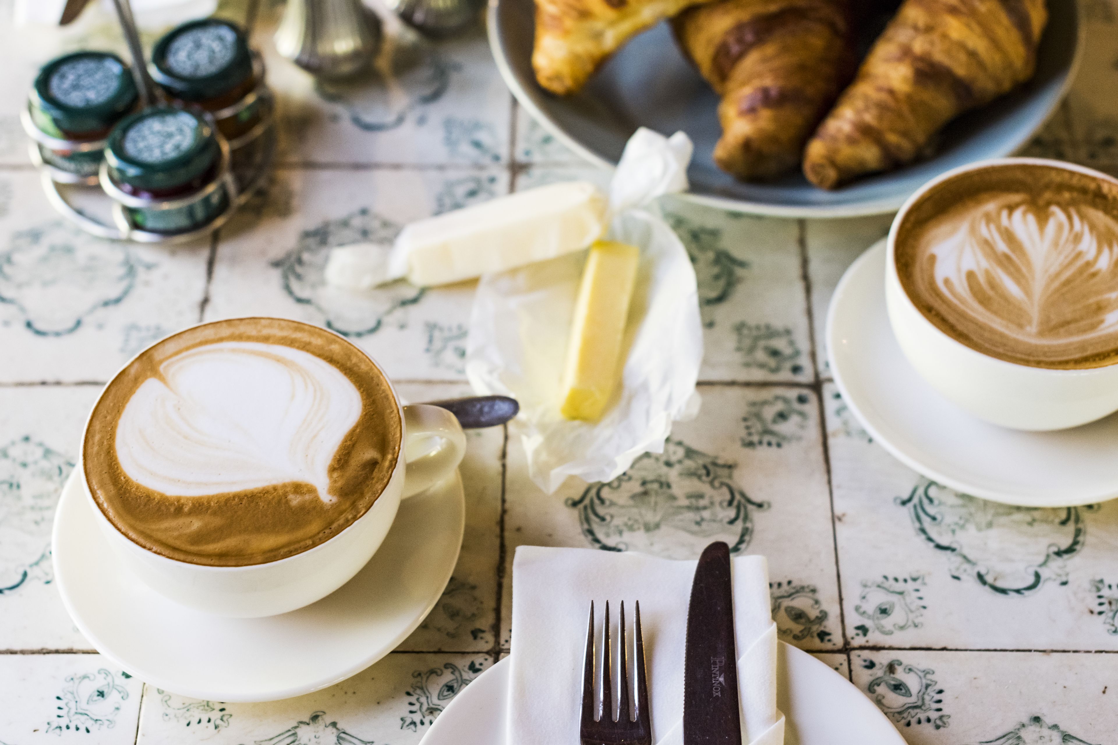 A table with a croissant, a croissant plate, a butter dish, a knife, a fork, and two cups of coffee.