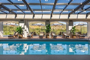 A pool area with lounge chairs and umbrellas, surrounded by a glass-enclosed structure with a view of the landscape. - Lanzerac Hotel (Stellenbosch)