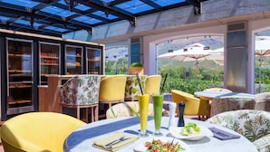 Un espace de restauration extérieur baigné de lumière, avec des tables rondes, des chaises jaunes et une structure en verre qui le protège.