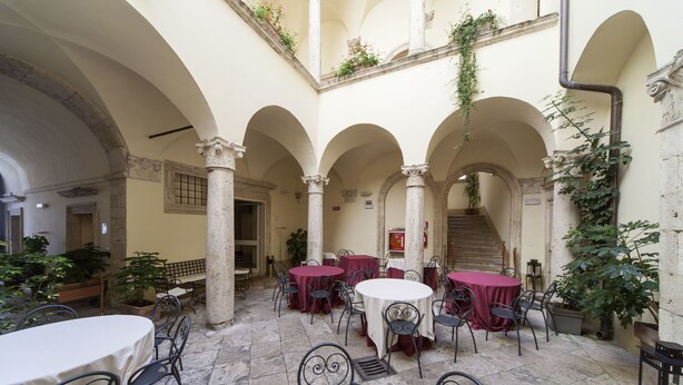 An indoor courtyard with tables and chairs, stone columns, and potted plants. - Palazzo Guiderocchi (Ascoli Piceno)