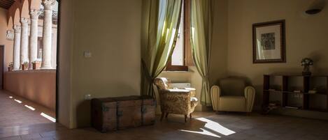 Una habitación con suelo de madera, una maleta antigua, un sillón estampado y una ventana con cortinas verdes.