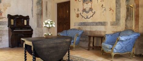 Una habitación con una mesa de madera oscura, sillas tapizadas en azul, un piano de madera y una pared decorada con frescos.