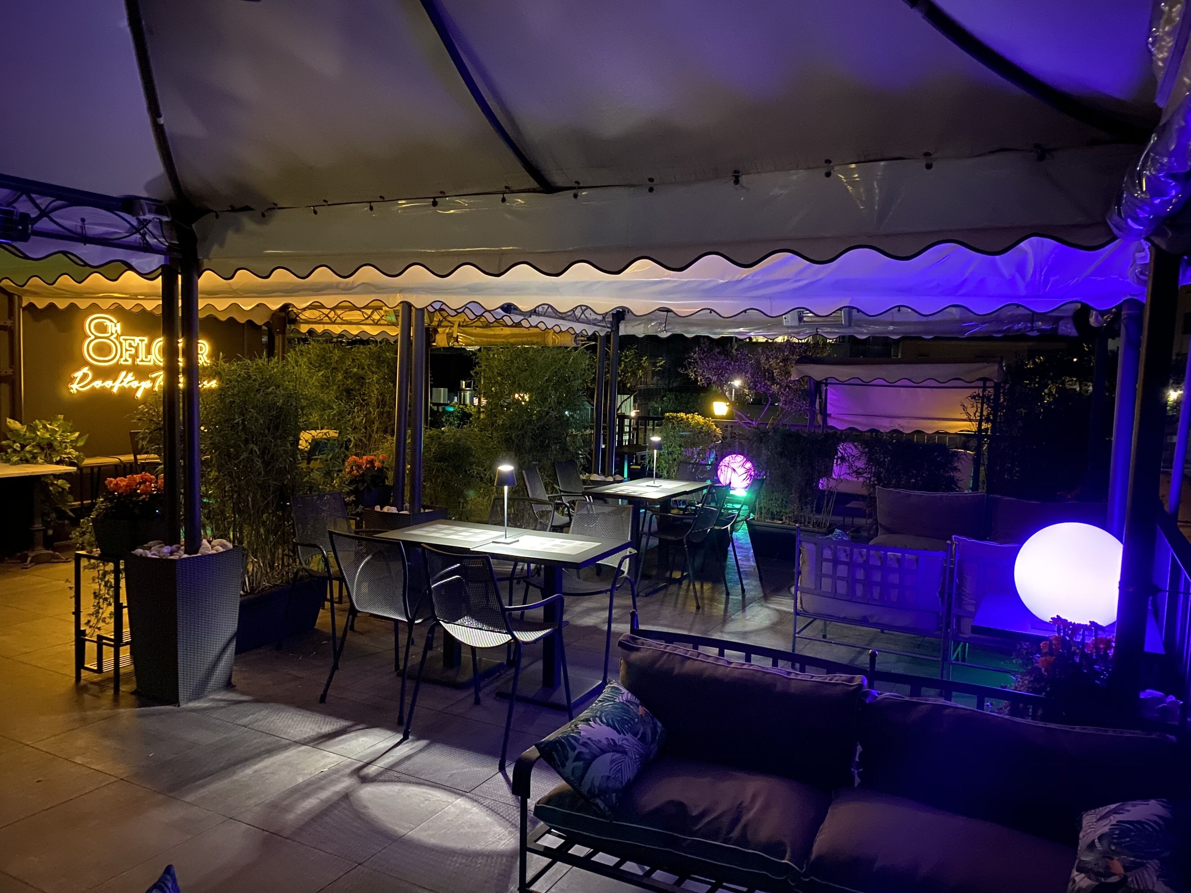 Ein Außenbereich mit einer Terrasse, Tischen, Stühlen und einer Sitzecke, sowie einem Neon-Schild.
