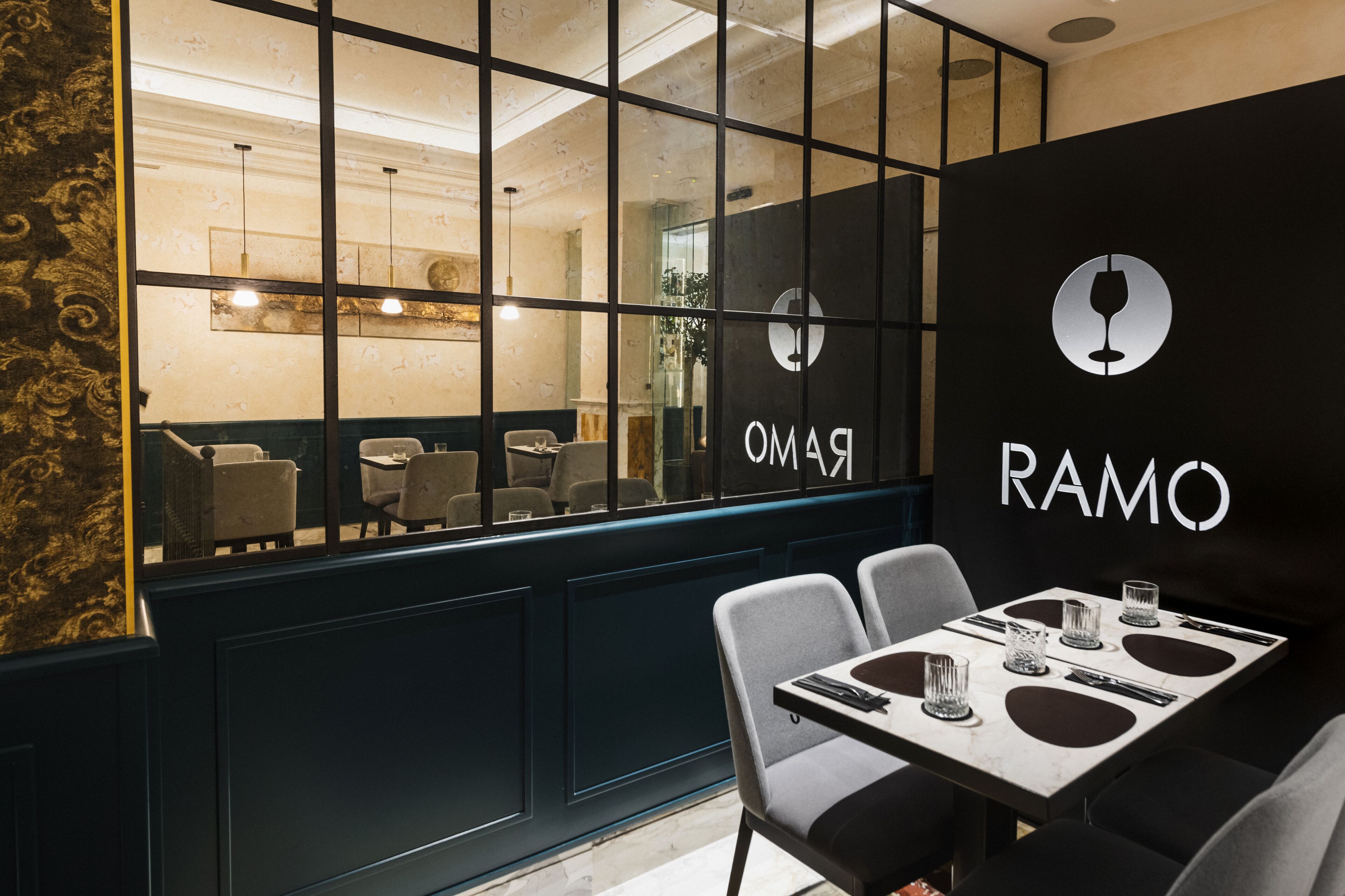 Et restaurantinteriør med et moderne design, der inkluderer en sort væg med logoet "RAMO" og en glasvæg med udsigt til spiseområdet.