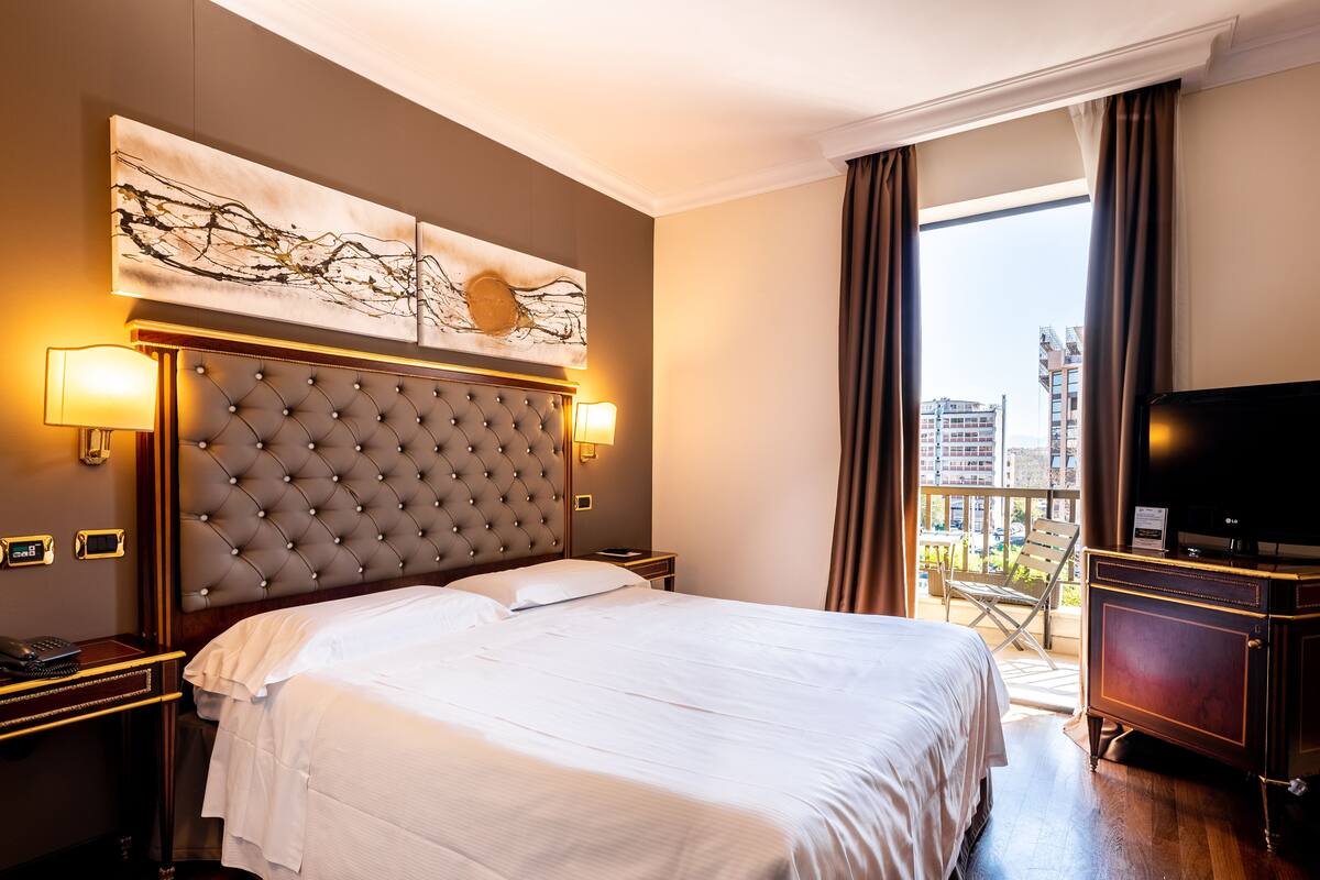 A hotel room with a tufted headboard, a large bed, a flat-screen TV, a balcony with a view of the city, and a painting on the wall.