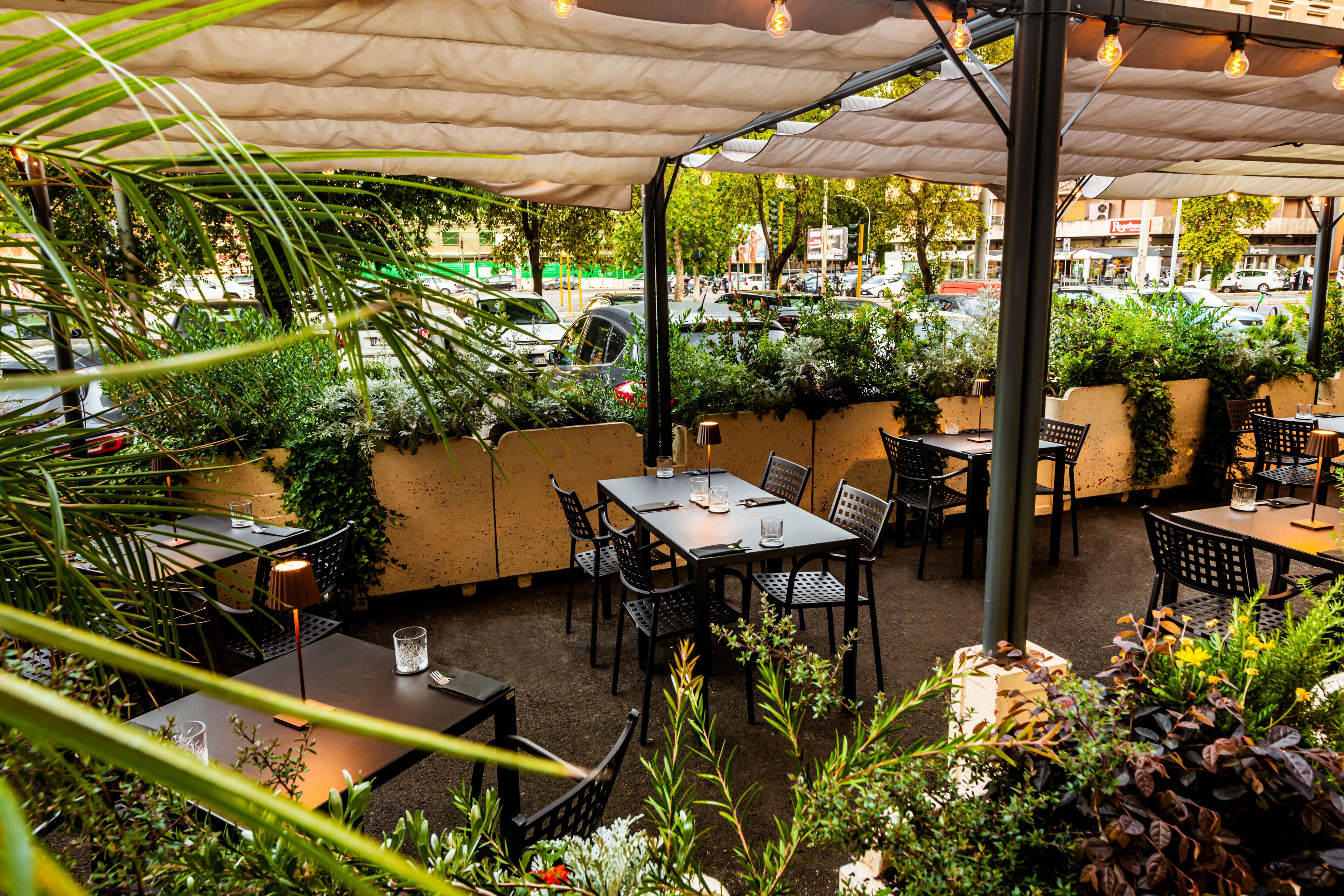 En udendørs terrasse med borde og stole, omgivet af grønt og udsigt til gaden.