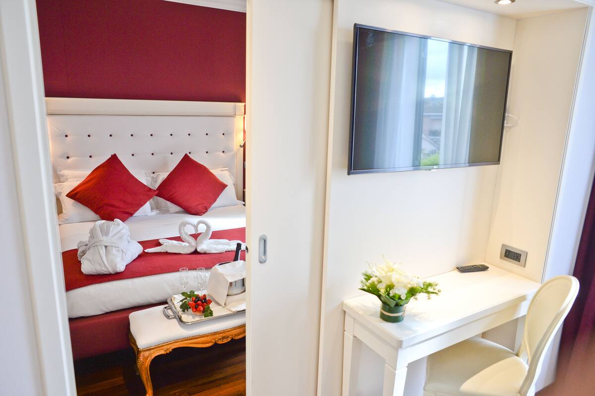 A hotel room with a bed, a desk, a chair, a TV, and a small table with a tray of fruit and a vase of flowers.