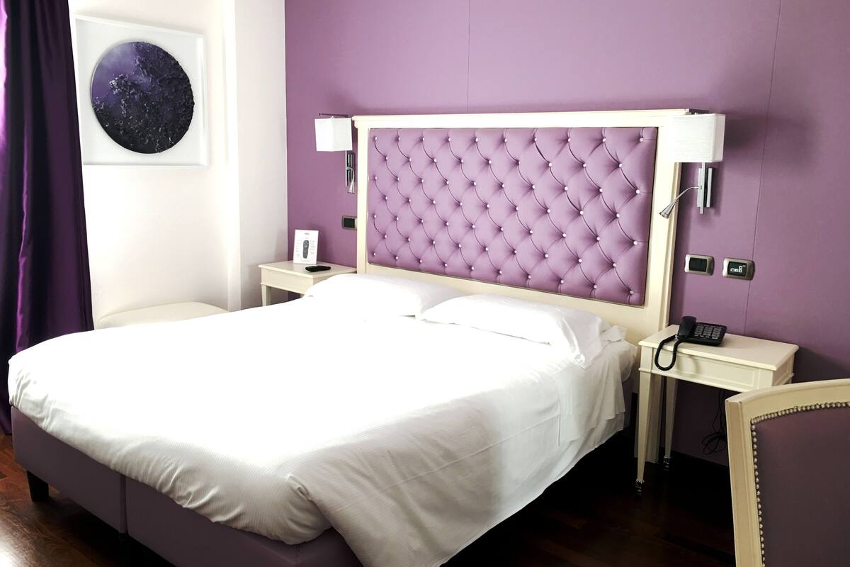 A neatly made bed in a room with a purple headboard, white bedding, and a small bedside table with a lamp and a phone.