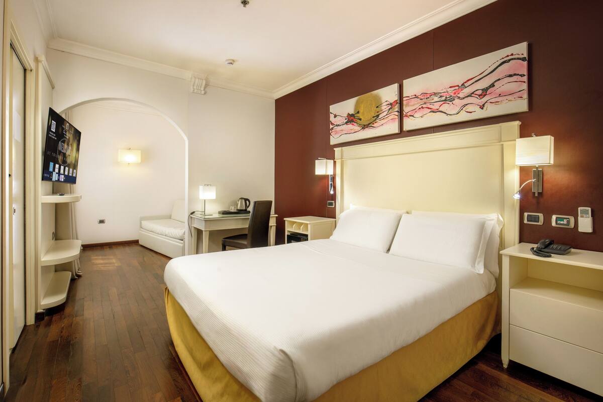 A hotel room with a large bed, a desk, a chair, a lamp, and a painting on the wall.
