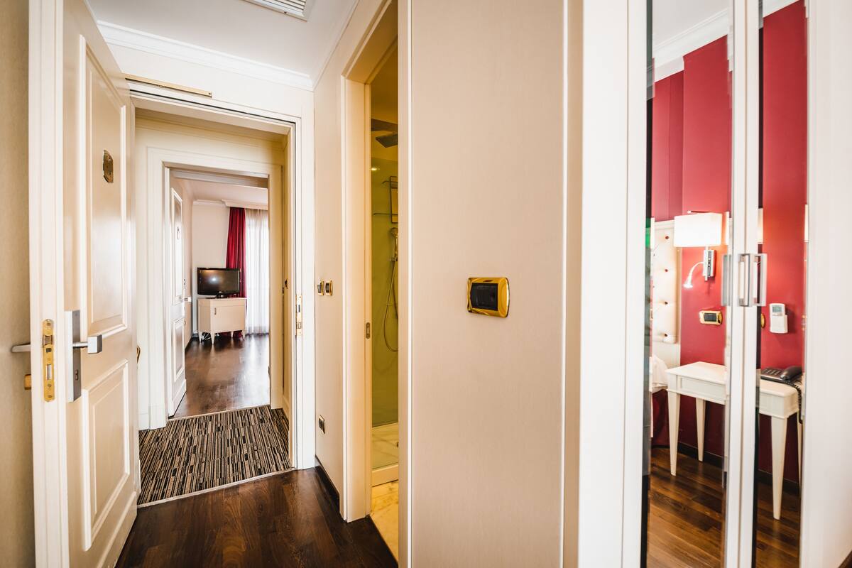 A hotel corridor with a wooden floor, a red wall, and a white door leading to a bathroom with a shower and a toilet.