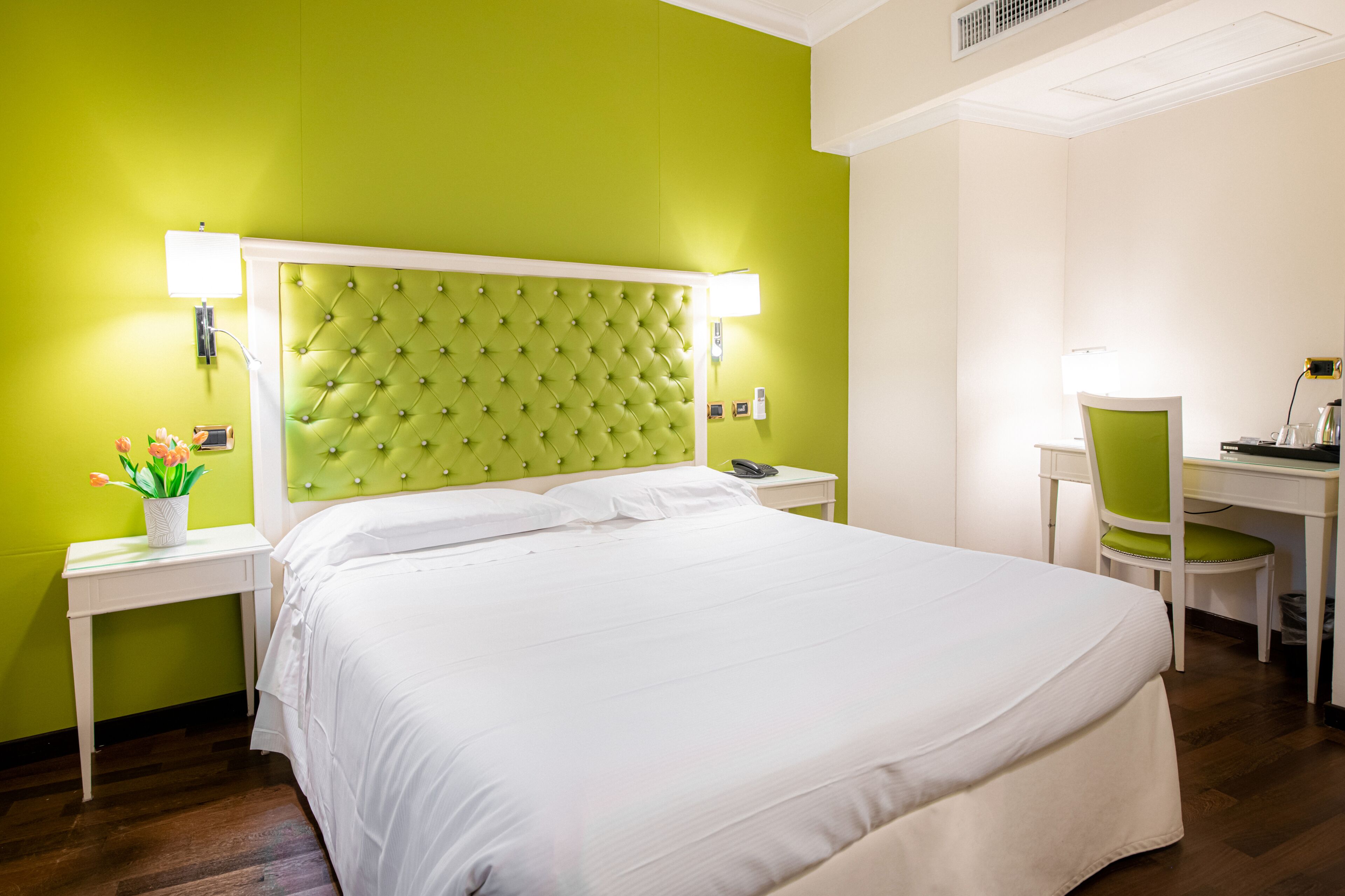Una camera d'albergo con una testiera verde, biancheria da letto bianca, una scrivania con una sedia e un piccolo tavolino con una pianta.