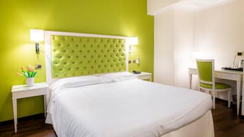 Una camera d'albergo con una testiera verde, biancheria da letto bianca, una scrivania con una sedia e un piccolo tavolino con una pianta.