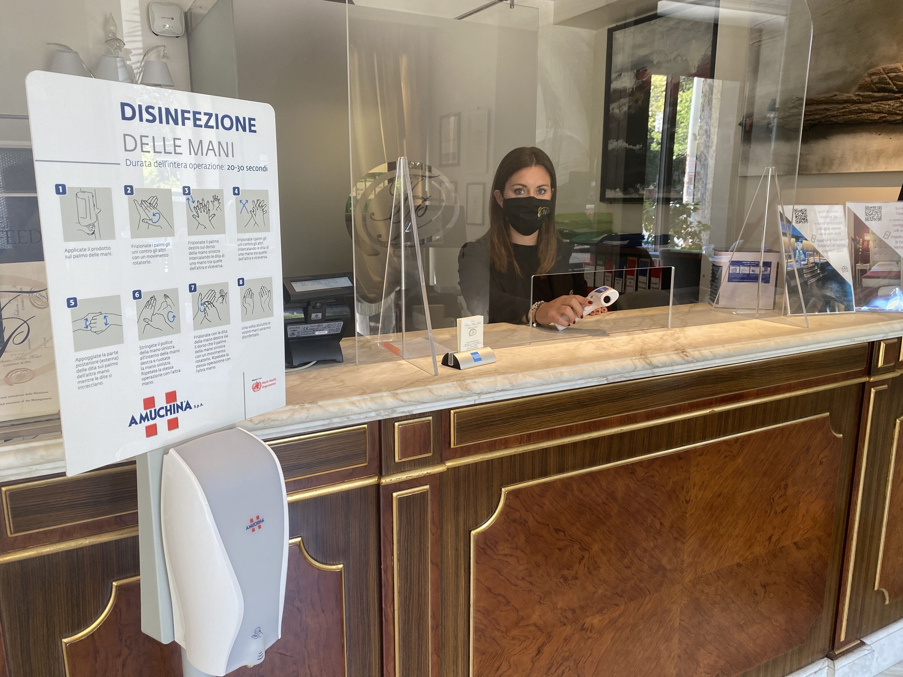 Ein Empfangstresen mit Handdesinfektionsmittelspender, einem Schild mit Anweisungen zur Handdesinfektion und einer Frau hinter dem Schalter.