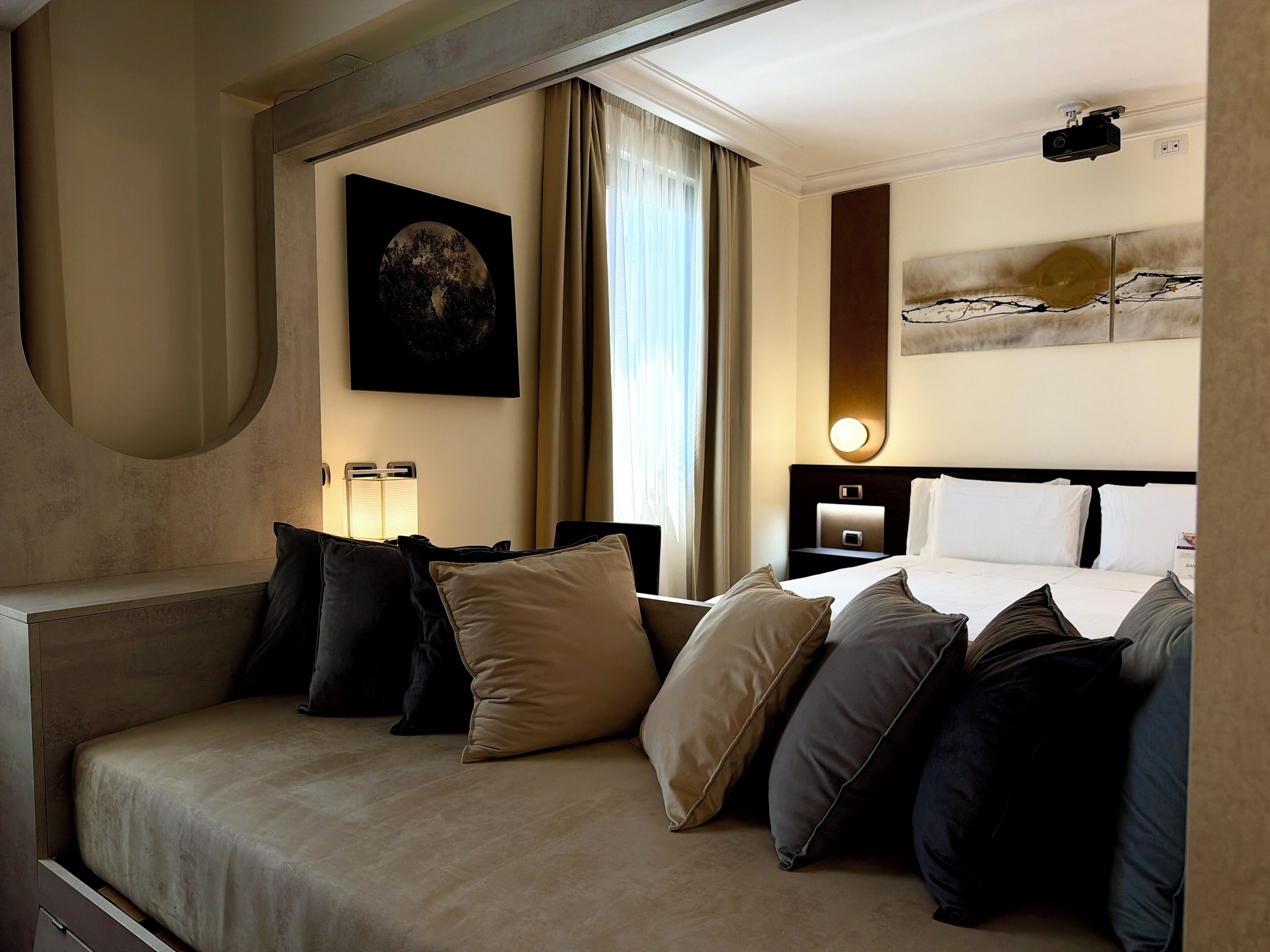 Habitación de hotel con sofá, cama con almohadas, mesita de noche, lámpara, un cuadro y un proyector en el techo.